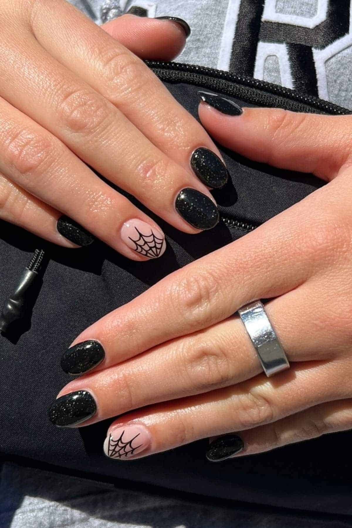 Hands with black manicured nails, two accent nails featuring a white base and black spiderweb design, holding a black pouch; person wears a silver ring.