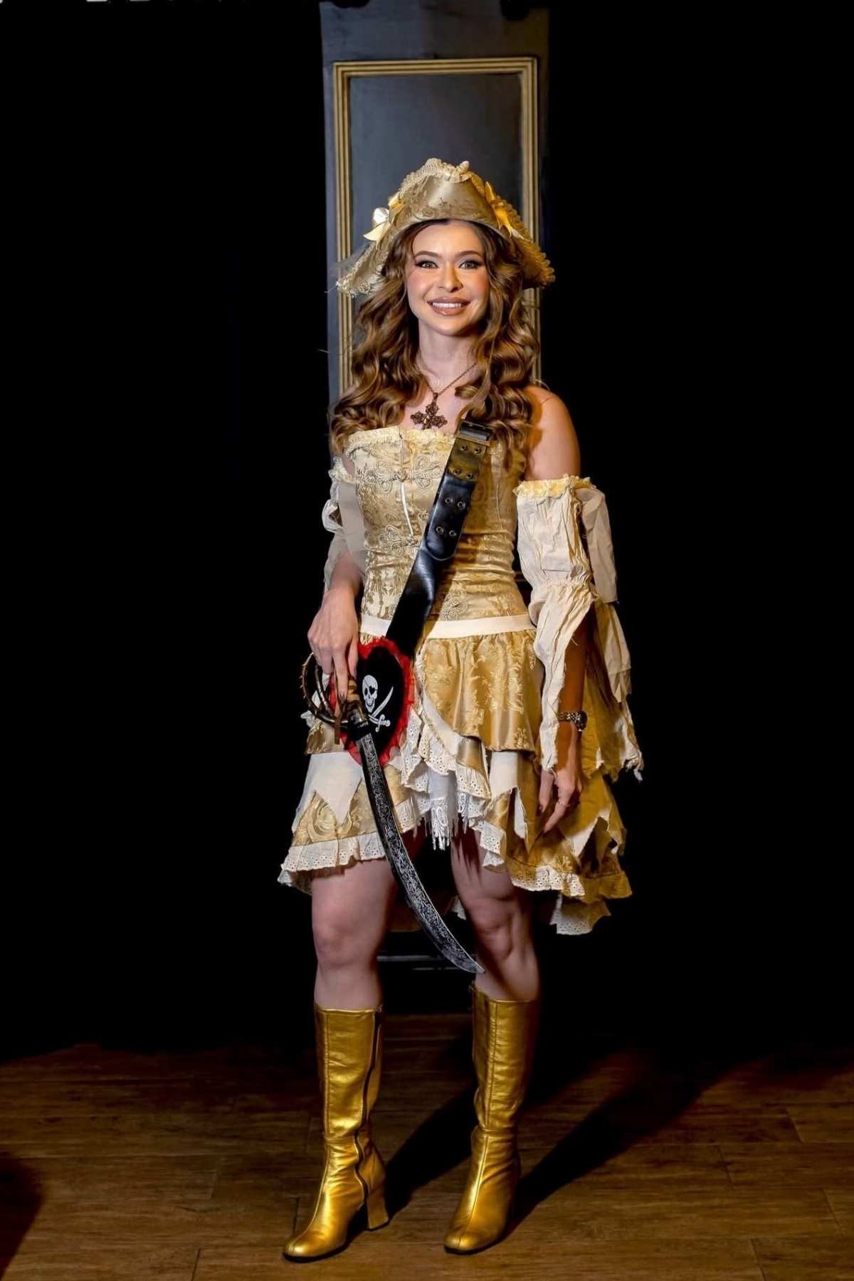 Woman in a gold pirate costume with a hat, sash, and gold boots stands and smiles against a dark background.