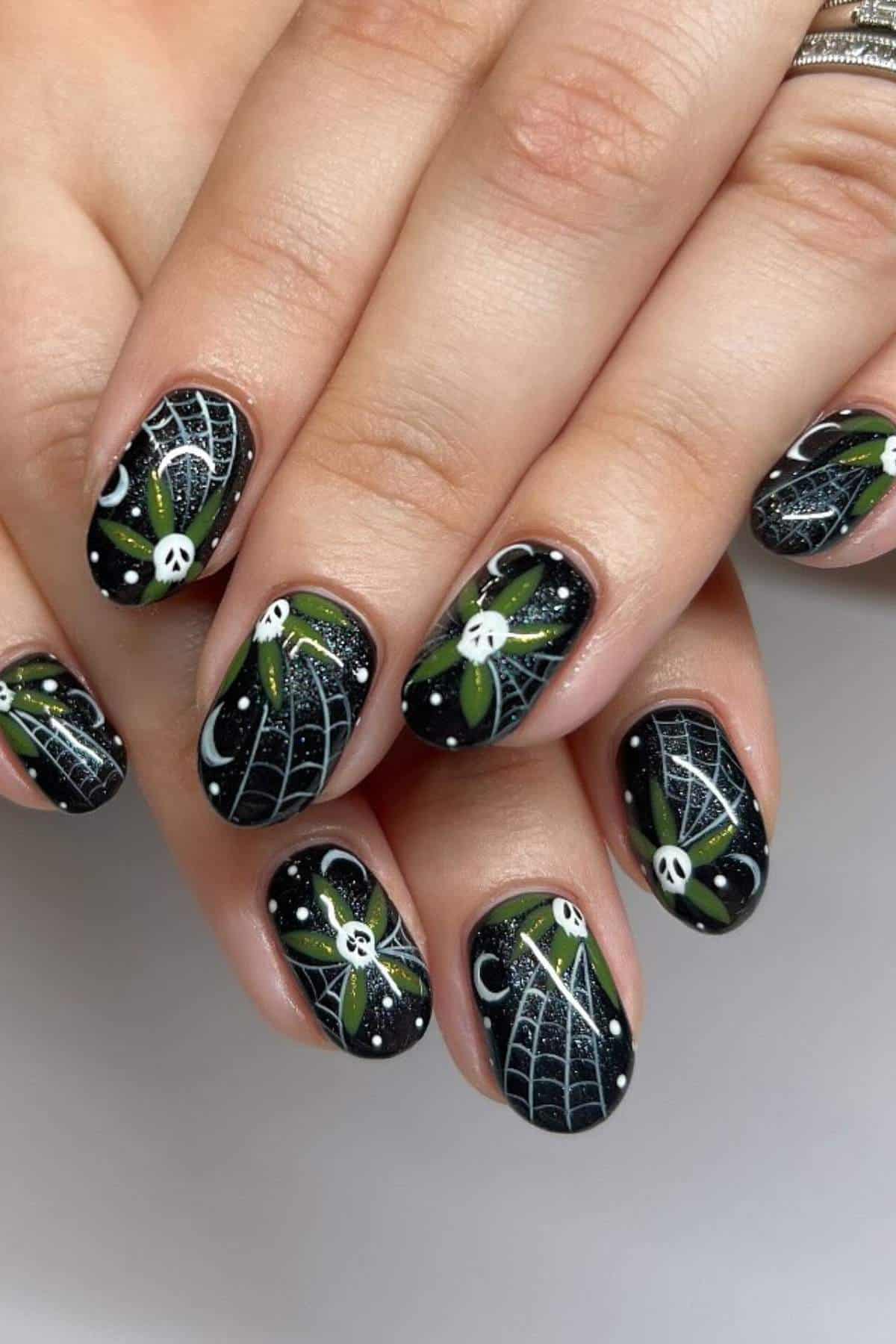 Close-up of fingernails with black polish, white spider web designs, small white faces, green leaves, and white crescent moons and dots; hands are gently overlapping.