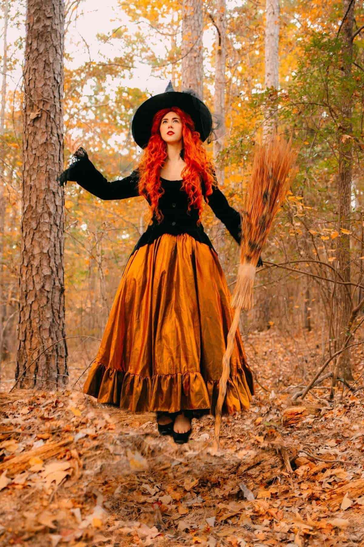 A woman with long red hair in a black hat and orange dress holds a broom while standing in a forest with autumn leaves.