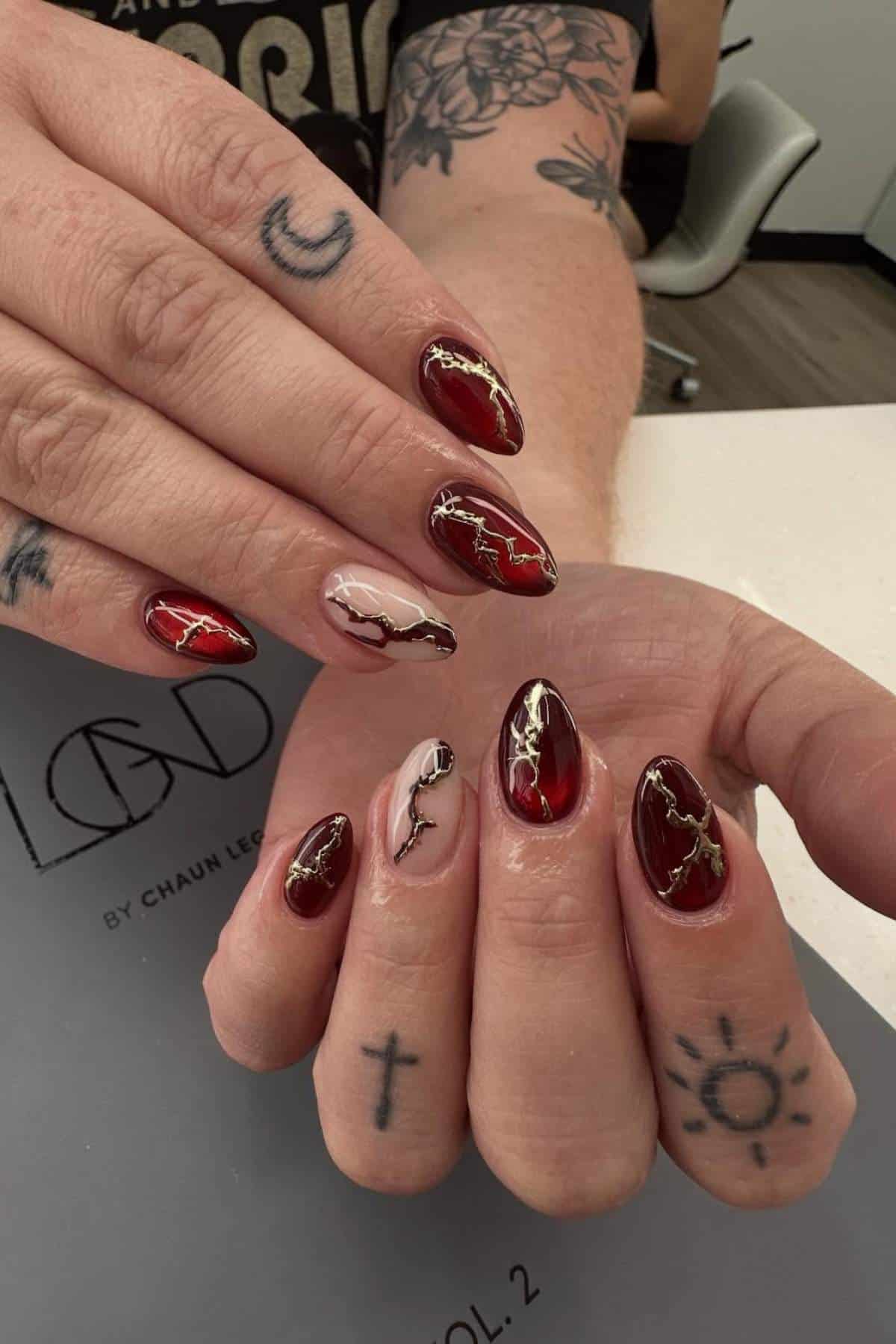 Hands with tattoos display almond-shaped nails featuring a red, nude, and gold marbled design. The background includes a nail salon setting with a gray mat on the table.