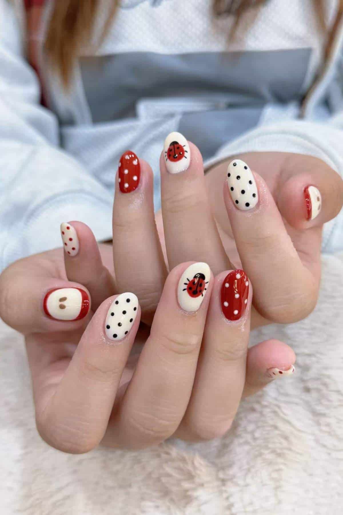 Hands with manicured nails featuring red, white, and black designs, including polka dots and ladybugs, against a light background.