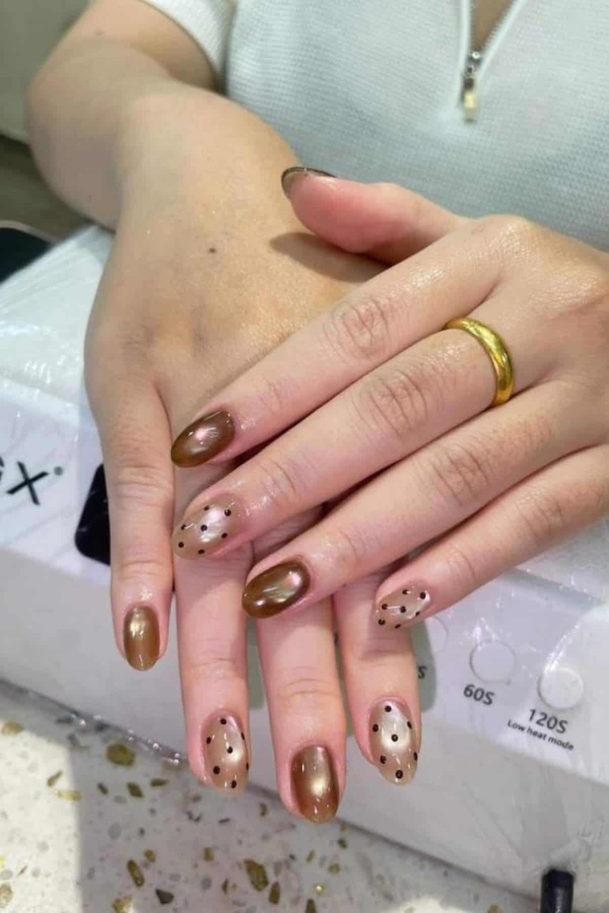 A person with almond-shaped nails featuring brown polish and black polka dot accents, wearing a gold ring on their ring finger.