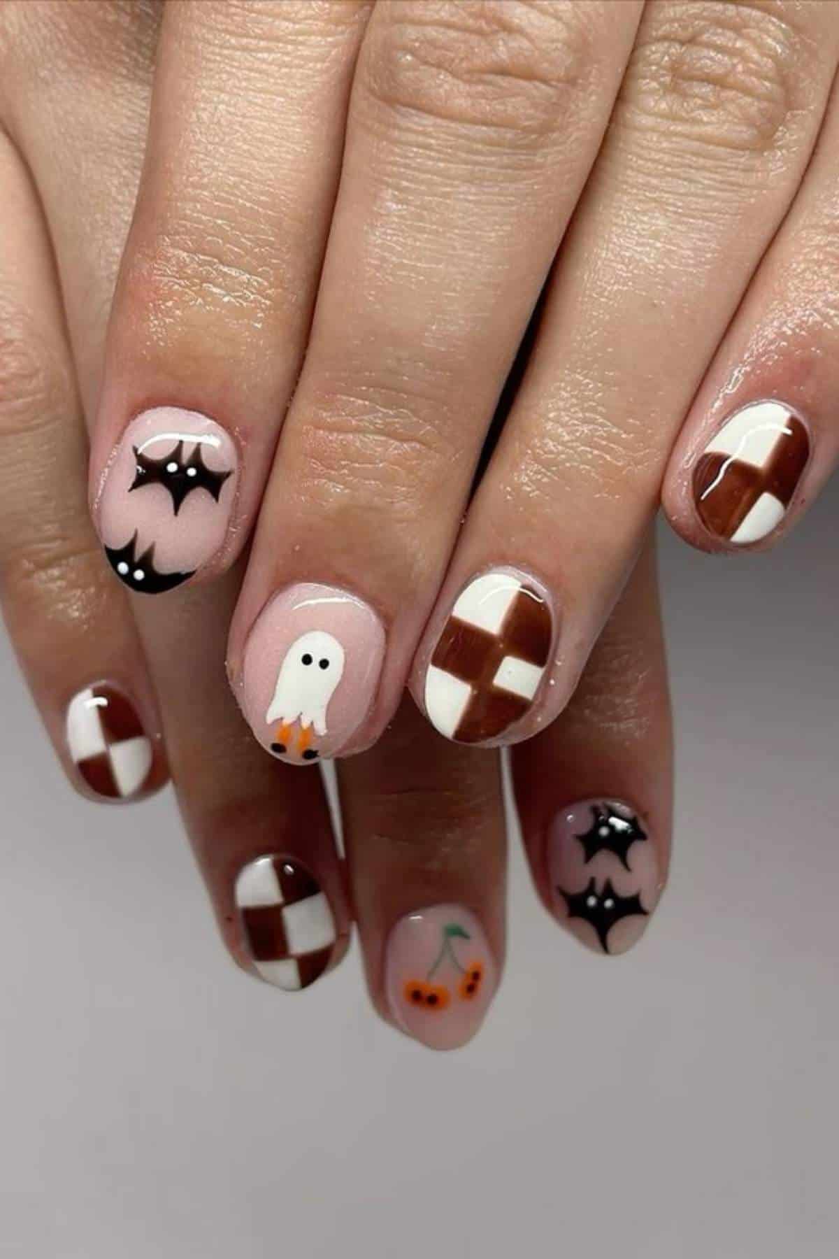 A hand with short nails painted with Halloween designs, including bats, a ghost, checkerboard patterns, and cherries in black, white, and brown polish.