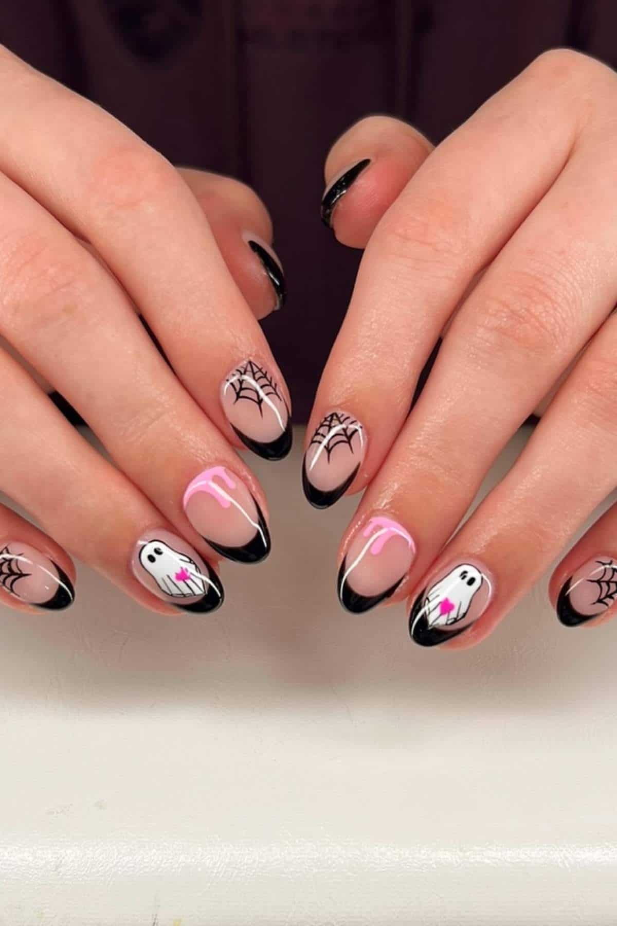 Hands with almond-shaped nails featuring black tips, spiderwebs, white ghosts, and pink drips on a light nude base, displayed against a white surface.