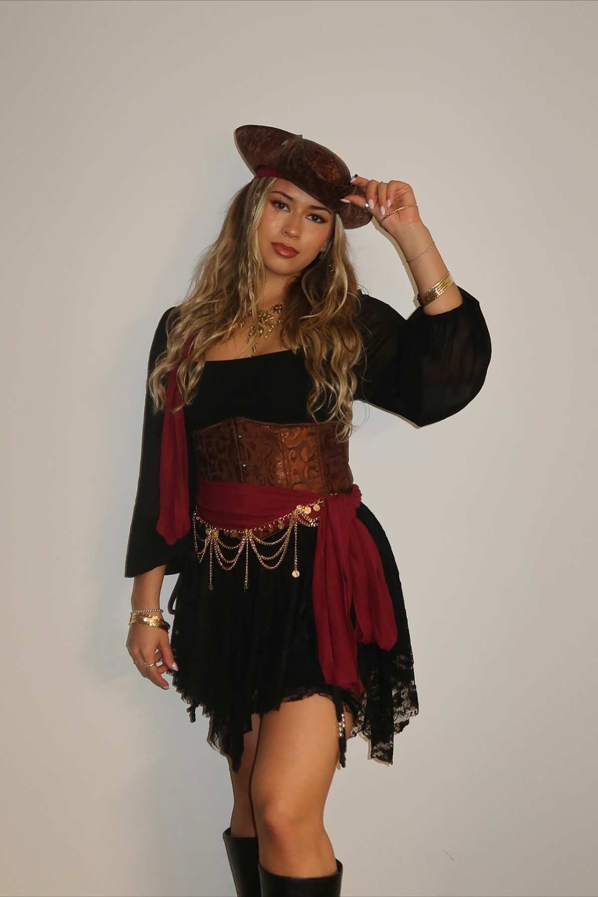 Woman poses in a pirate-inspired costume with a brown hat, black dress, red sash, gold jewelry, and knee-high black boots against a plain white background.