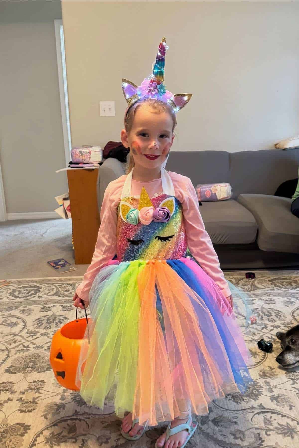A child in a rainbow unicorn costume holds a jack-o’-lantern bucket, standing on a patterned rug in a living room with a couch and toys in the background.