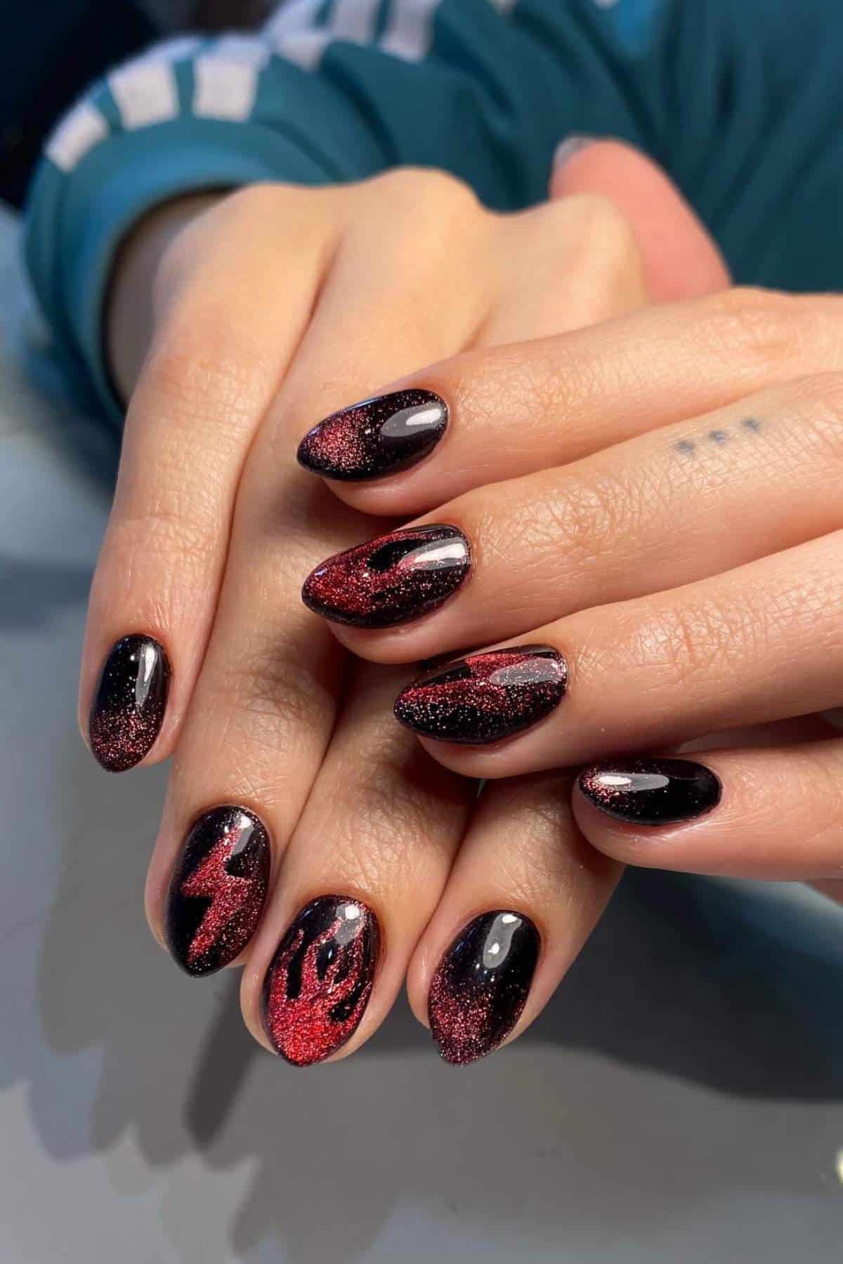 Hands with almond-shaped black nails featuring red glitter and abstract flame-like designs, resting on a flat surface.