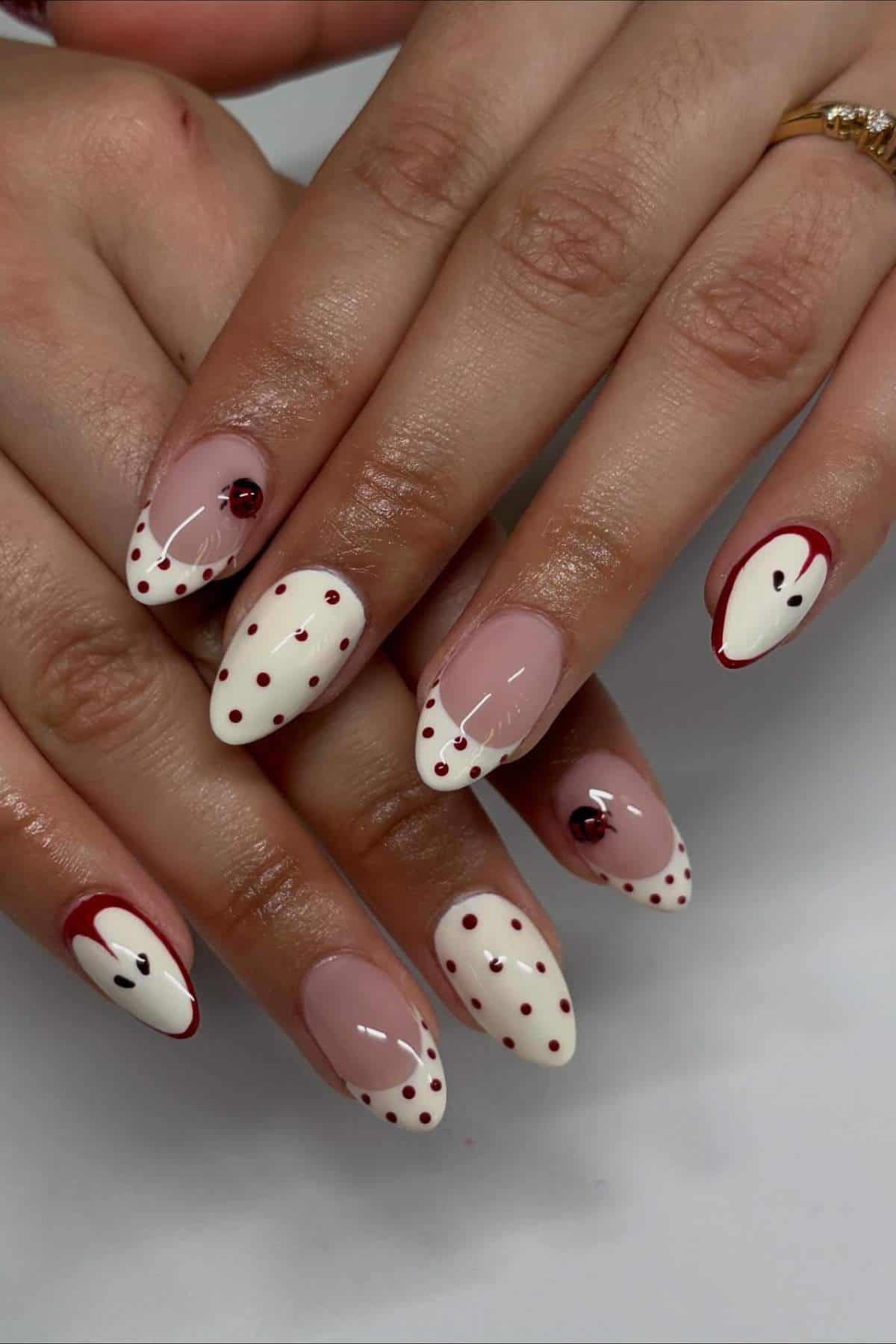 A hand with almond-shaped nails features a white French tip design with red dots and accents, some with red gems, on a natural pink base.