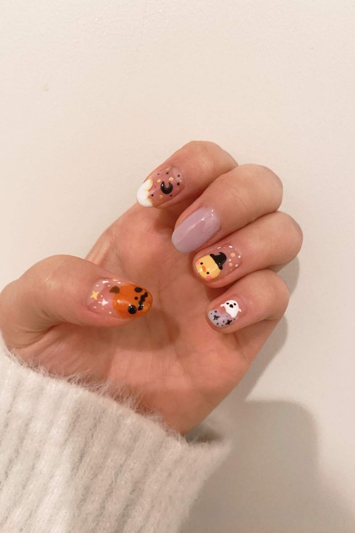 A hand with Halloween-themed nail art featuring a pumpkin, ghost, bat, cat, and polka dots on a light background.