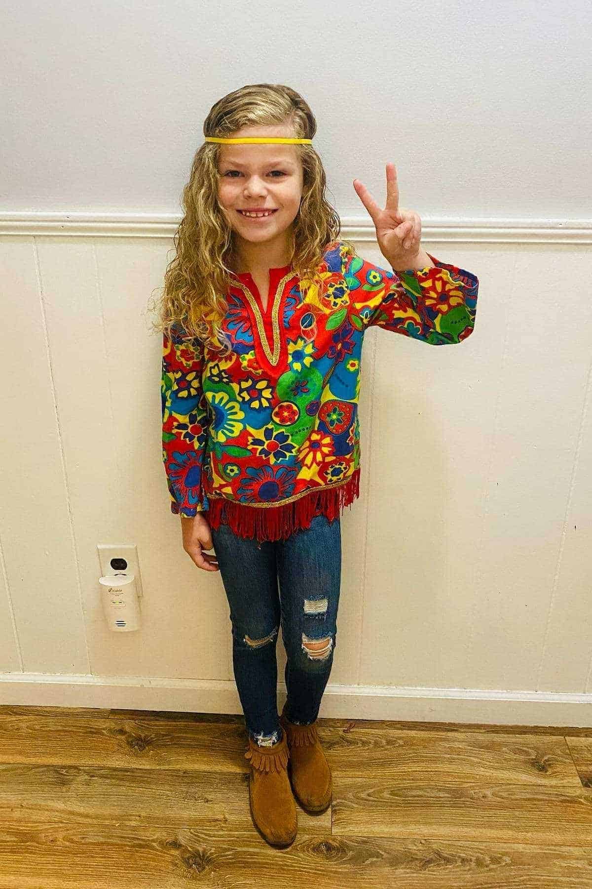 Young girl wearing a colorful floral shirt, blue jeans, brown boots, and a yellow headband, stands indoors making a peace sign with her right hand.