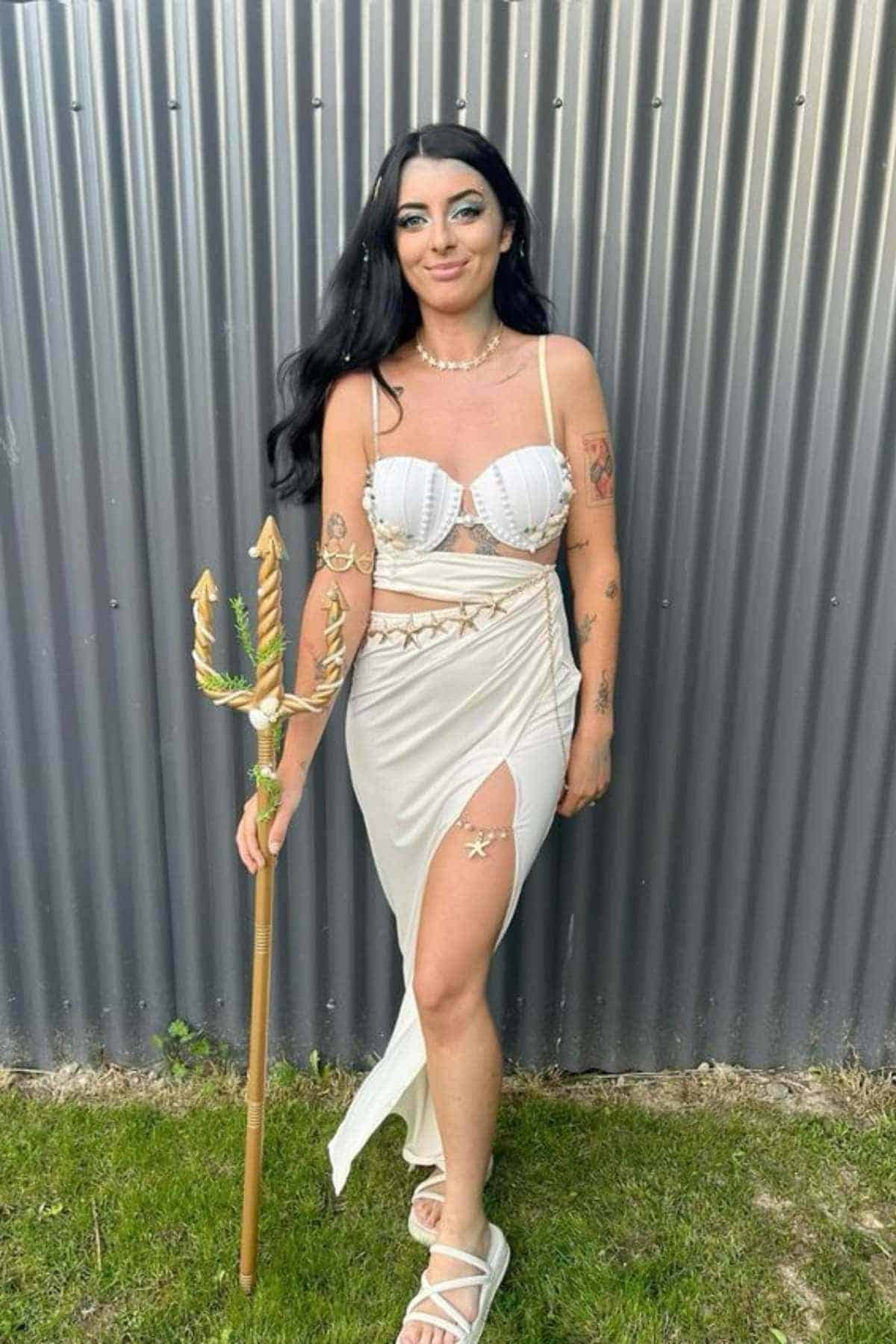 Woman in a white outfit holding a trident stands on grass in front of a corrugated metal wall.