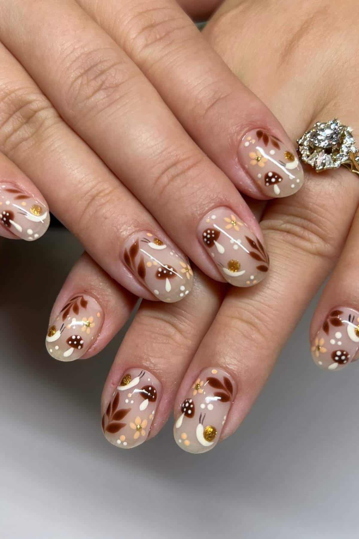 Hands with short, rounded nails featuring brown, beige, and gold floral and mushroom nail art designs; person wears a gold ring with a large gemstone.