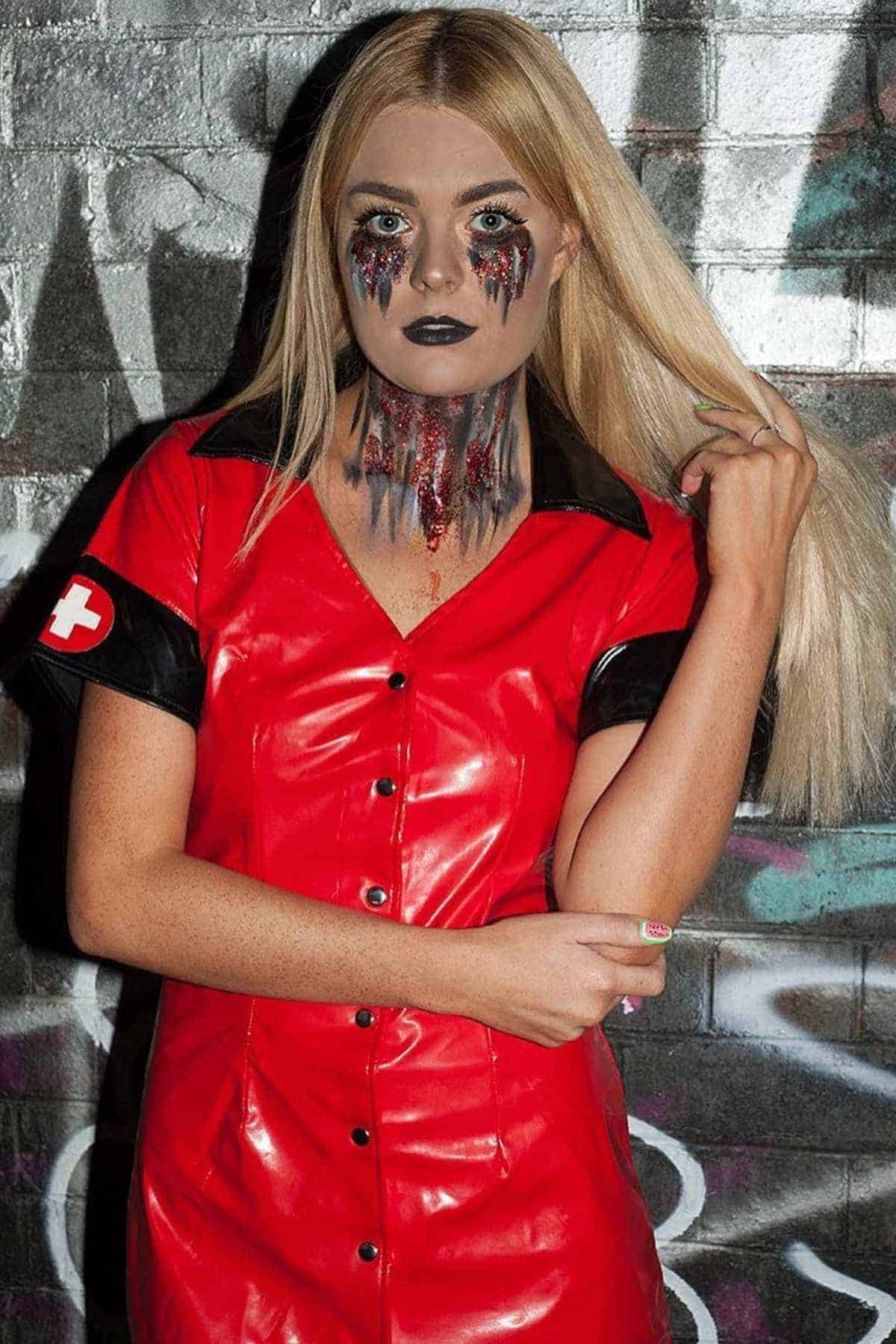 Person in a red nurse costume with black lipstick and dramatic makeup resembling melting black tears, standing in front of a graffiti-covered wall.