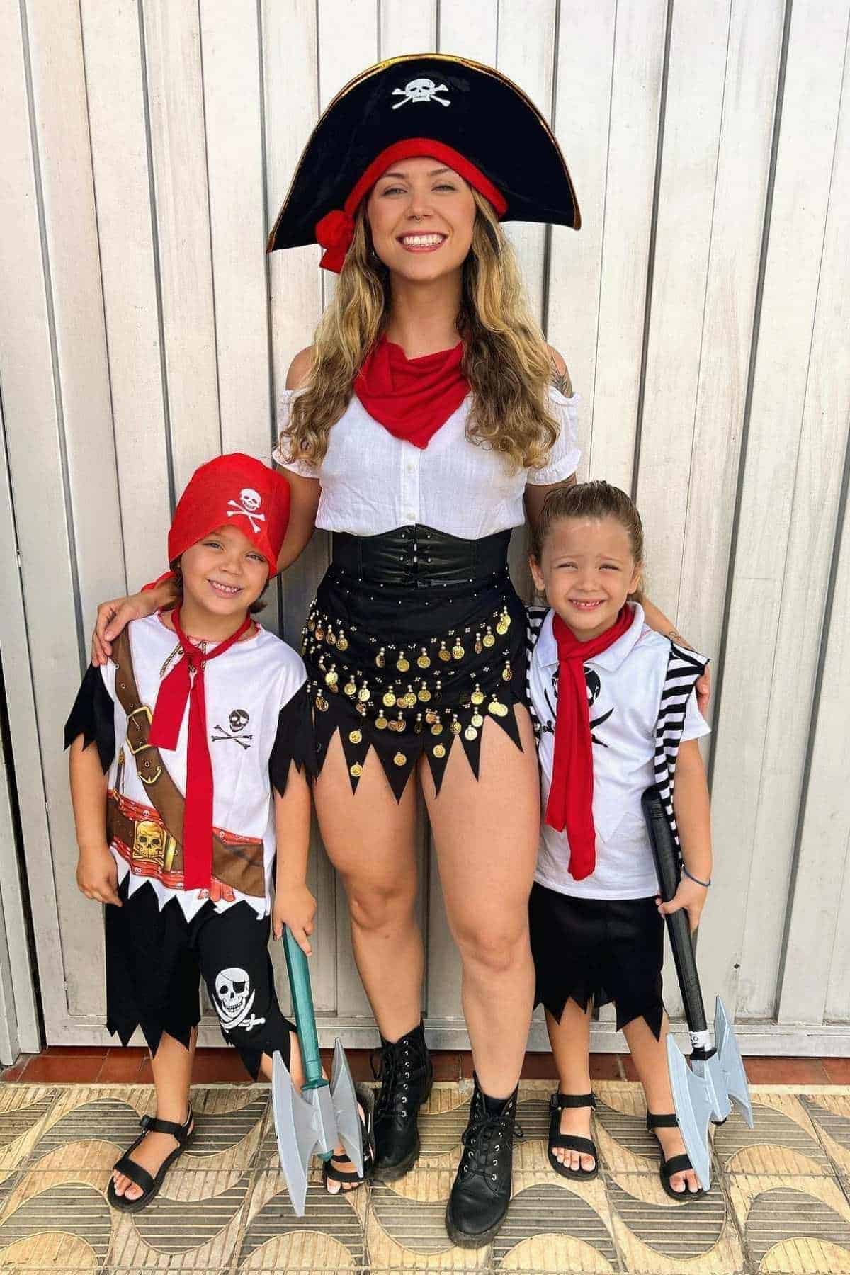 A woman and two children dressed as pirates stand smiling in front of a white wall; the kids hold toy axes and wear matching red bandanas.