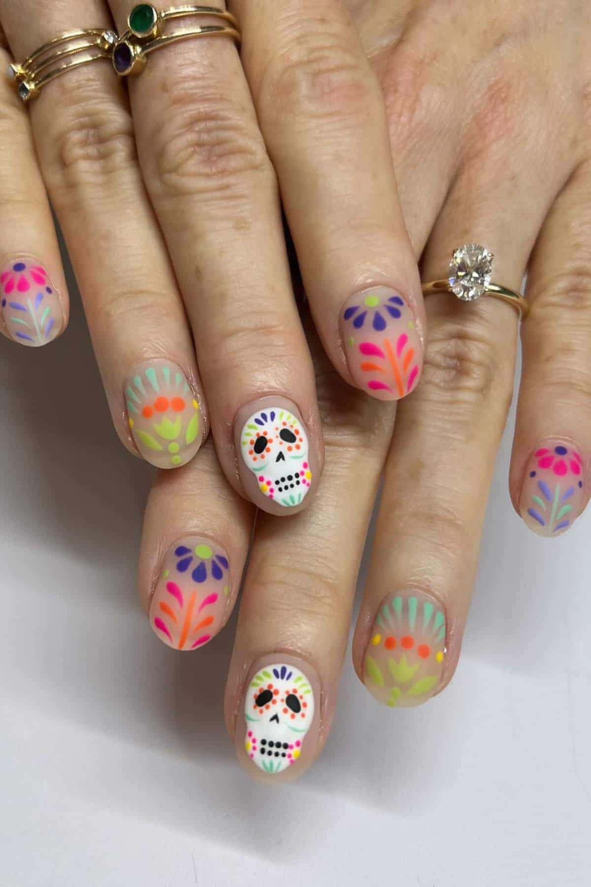 Hands with colorful, floral nail art and a sugar skull design on one nail. The person is wearing multiple gold rings, including one with a large clear gemstone.