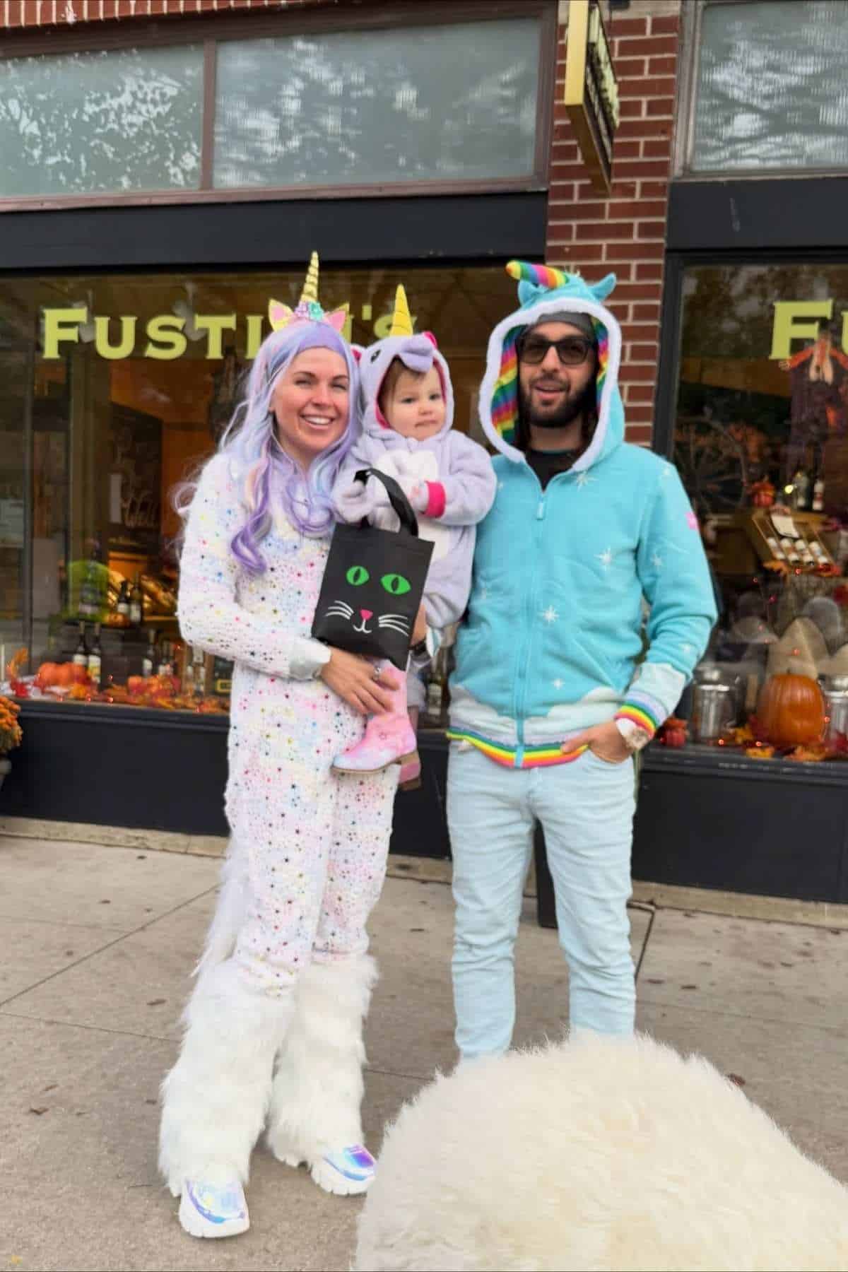 Three people in unicorn-themed costumes stand on a sidewalk outside a store. The adult on the left holds a child with a black cat bag. The adult on the right wears sunglasses.