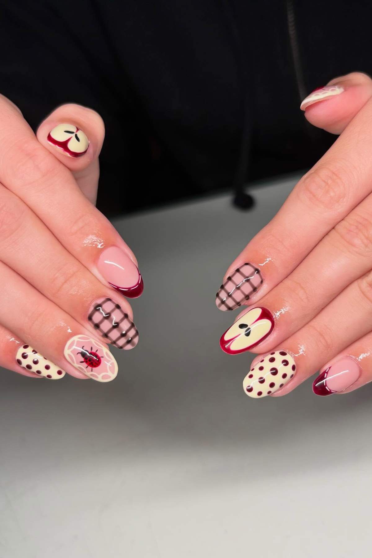 A pair of hands with manicured nails featuring red and beige designs, including apples, grids, dots, and a flower pattern.
