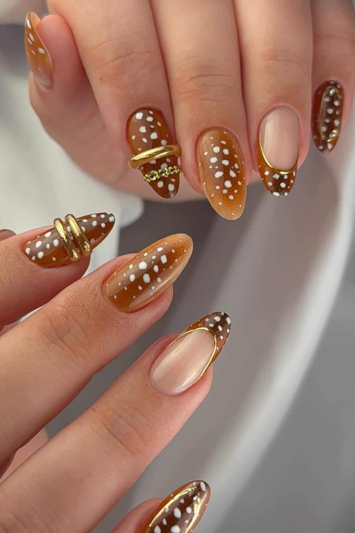 Close-up of hands with almond-shaped nails painted in brown ombre, white dots, gold accents, and gold rings on some nails.