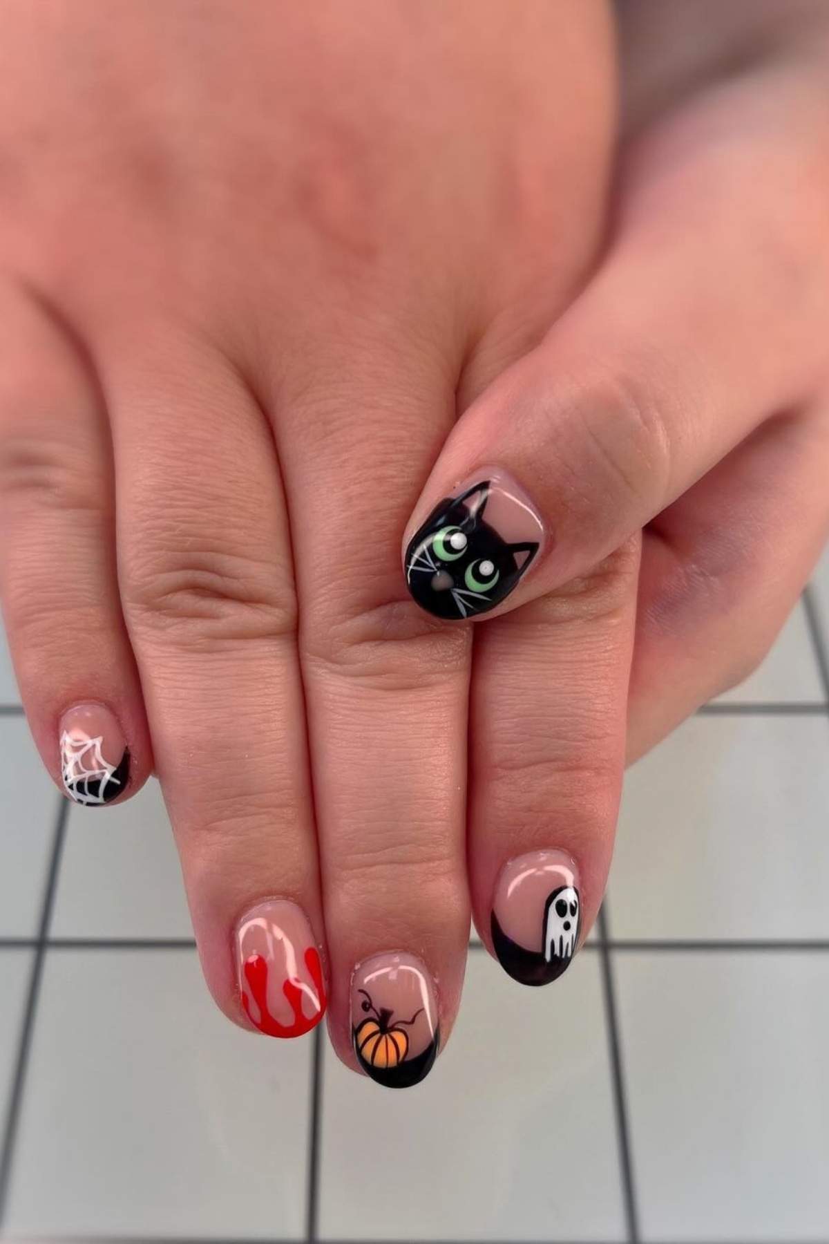 A hand with Halloween-themed nail art, including a cat, ghost, pumpkin, spiderweb, and red dripping design, on a tiled surface.