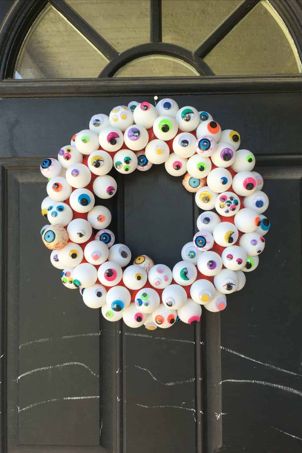 A wreath made of white balls painted with colorful eyeballs is hung on a black door.