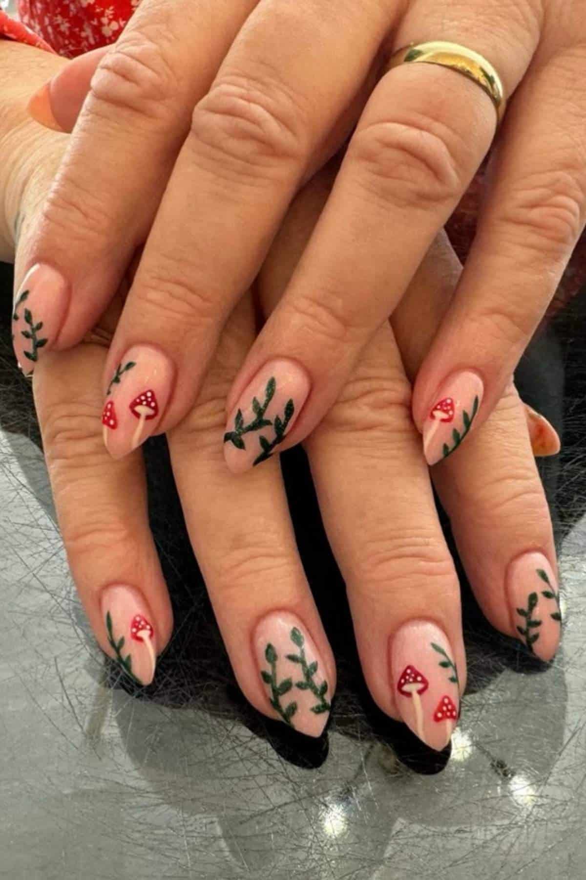 Hands with nude-colored nail polish featuring red mushroom and green leaf designs on long, almond-shaped nails. A gold ring is worn on the ring finger.