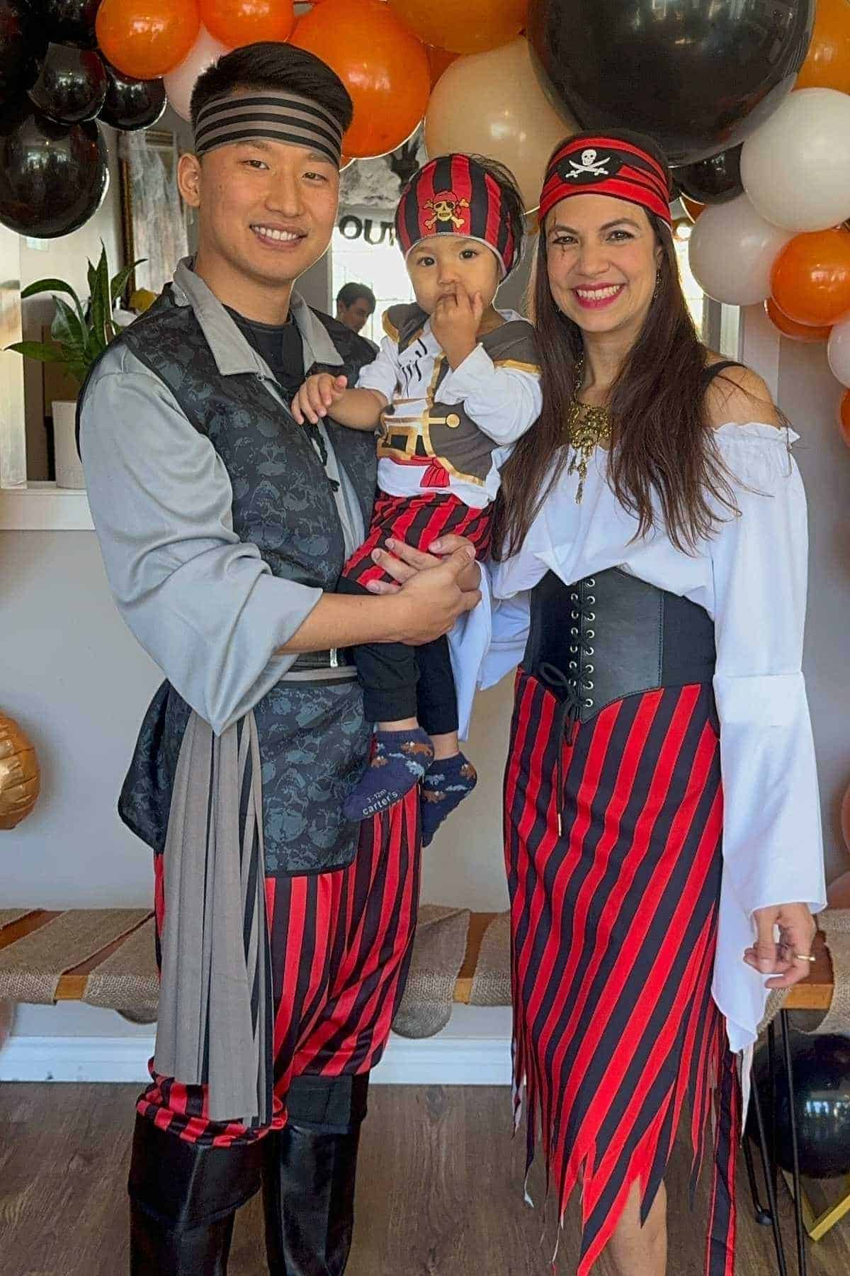 Three people dressed in pirate costumes stand together at a party decorated with black, orange, and white balloons.