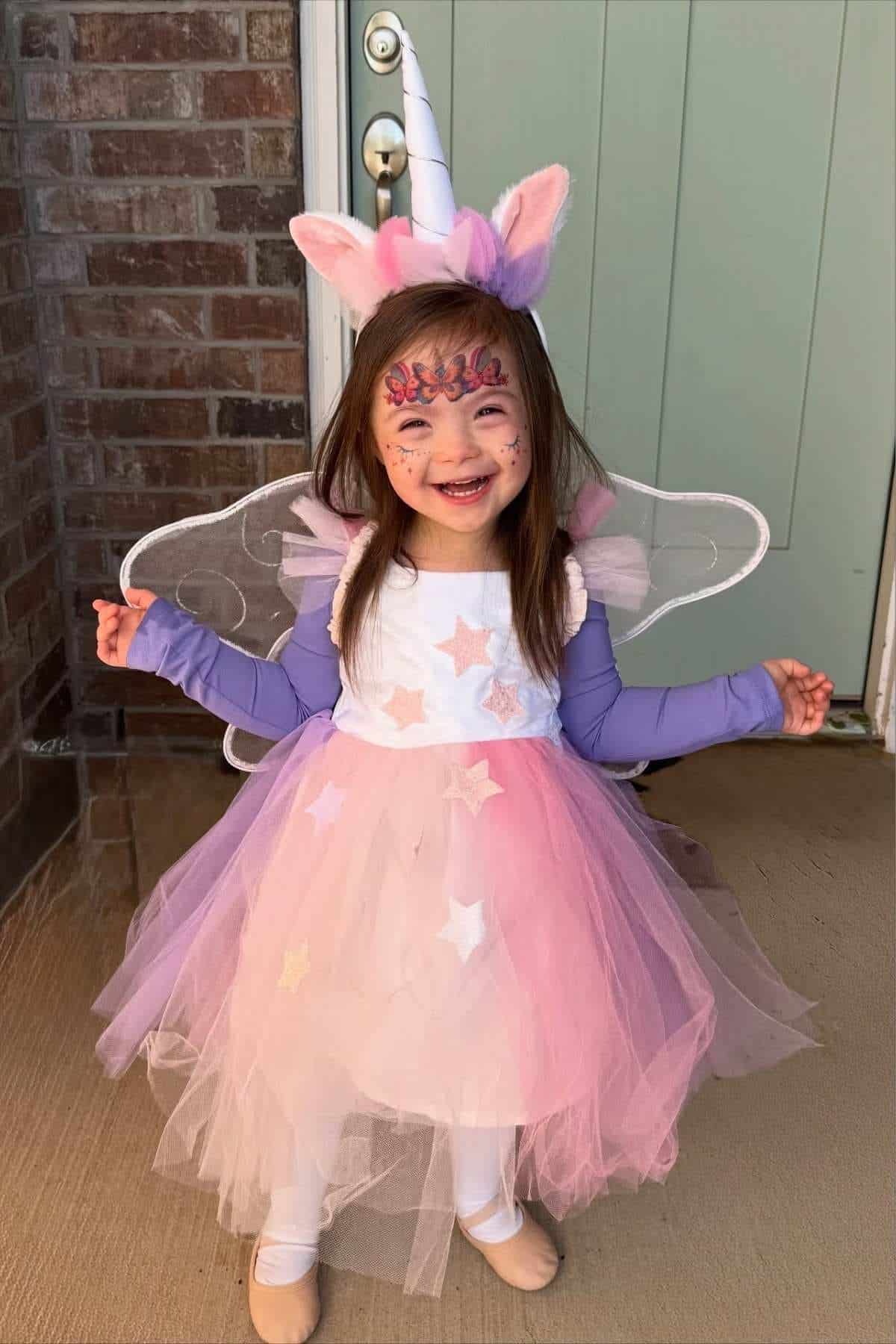 Young child in a unicorn costume with wings and face paint stands smiling on a porch in front of a green door and brick wall.