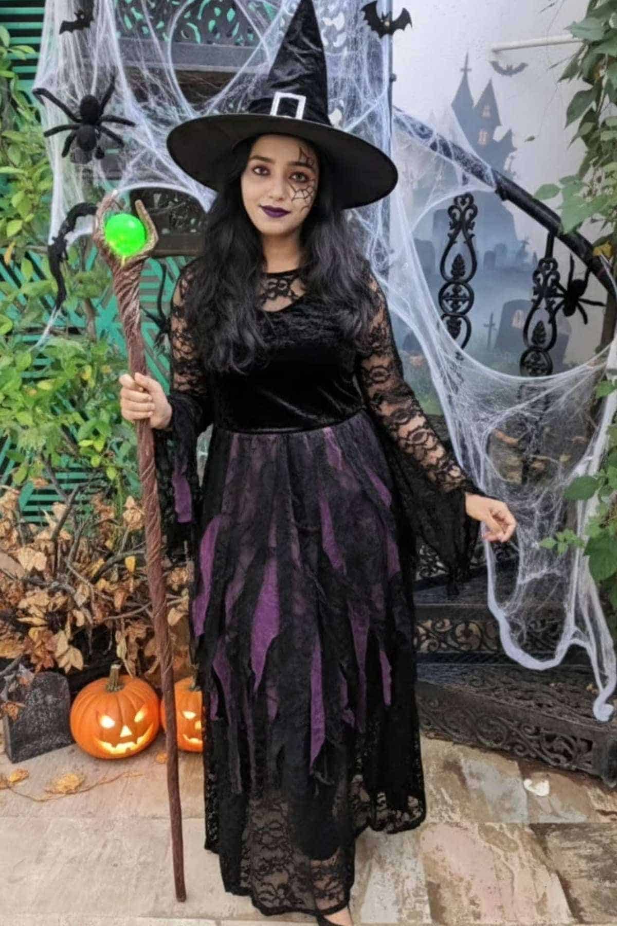Person in a black and purple witch costume with a hat and staff stands by pumpkins and Halloween decorations, including spider webs and a haunted house backdrop.