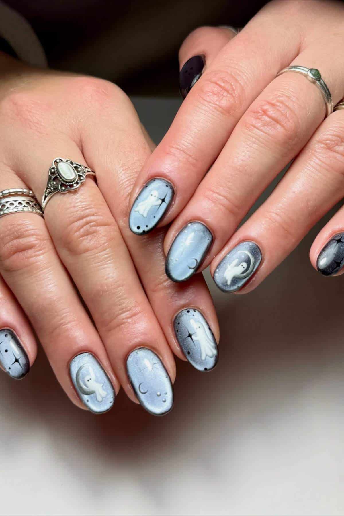 Hands with silver rings and pastel blue nail art featuring white ghost designs, small stars, and crescent moons.