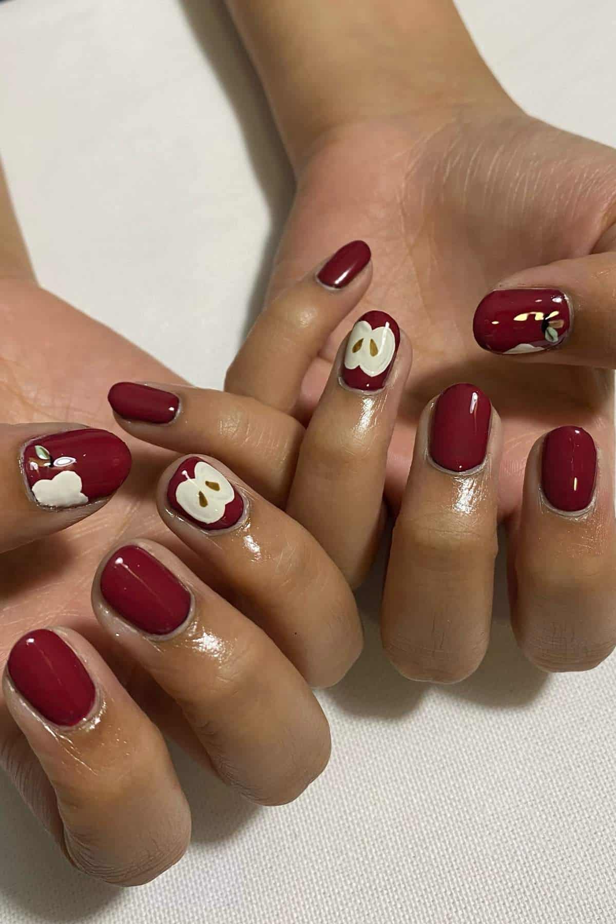 Fingernails painted dark red with accent nails featuring apple and apple slice designs on a white background.