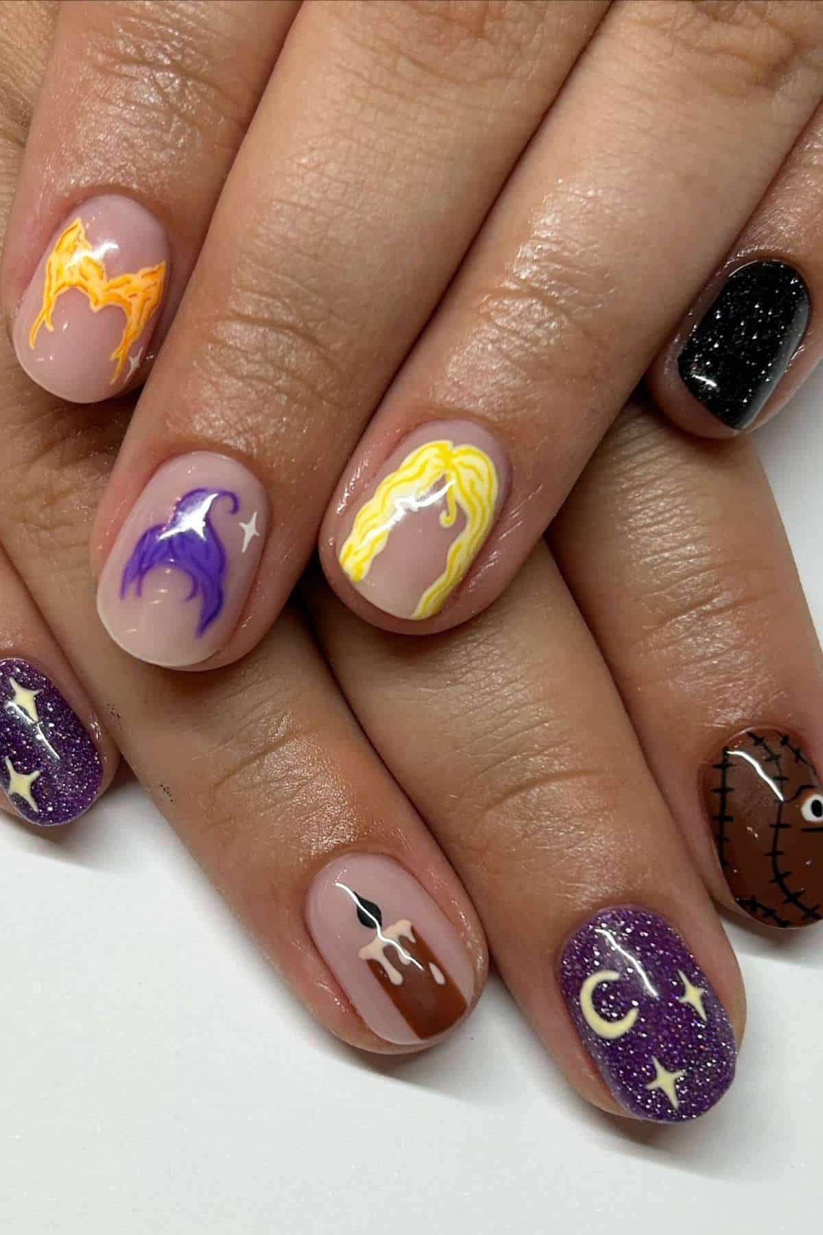 Hands with manicured nails featuring Halloween-themed nail art, including witches, moons, stars, stitches, and sparkling black and purple polish.