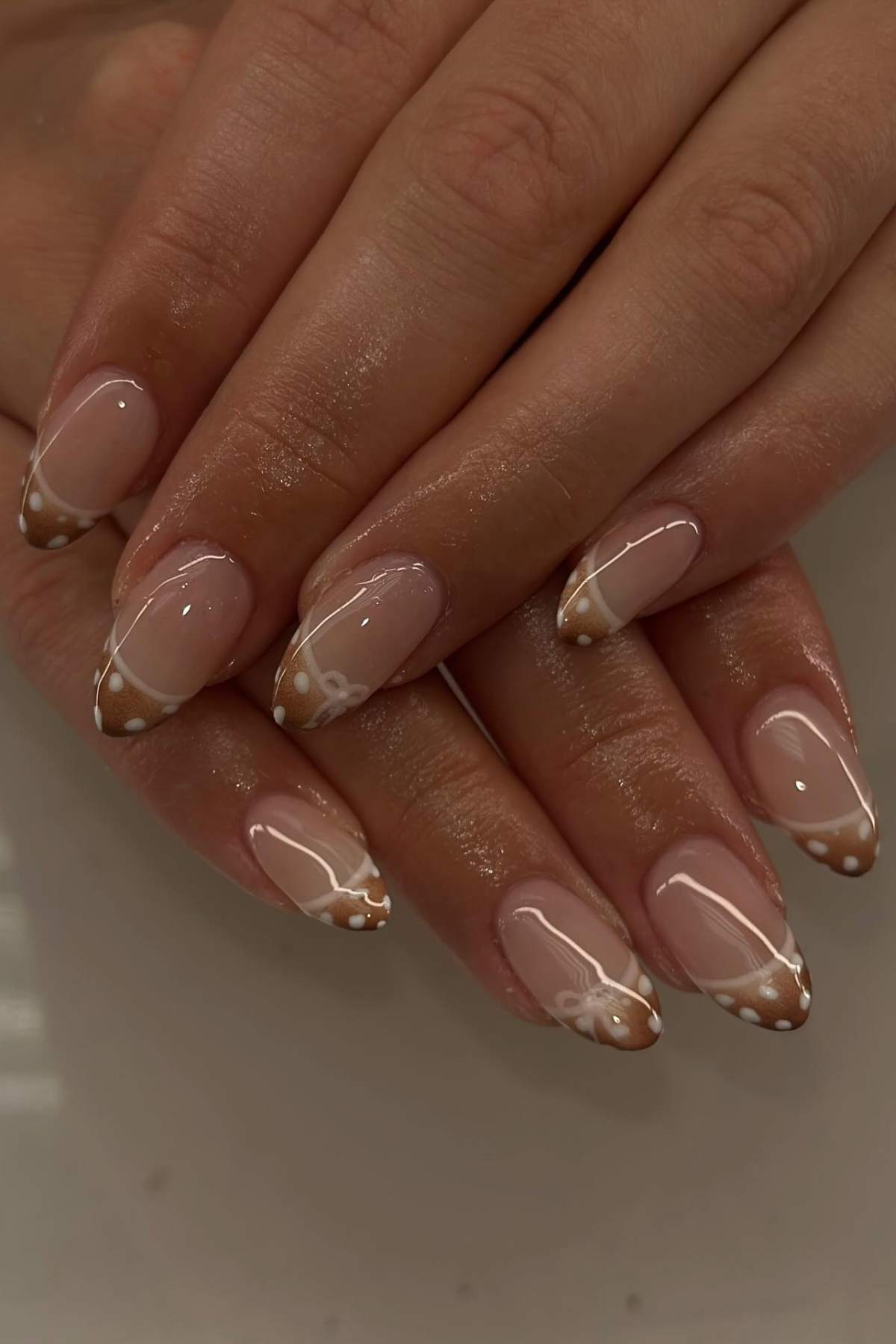 Hands with almond-shaped nails featuring nude polish, brown and white French tips, white dot accents, and small white bow designs on a few nails.