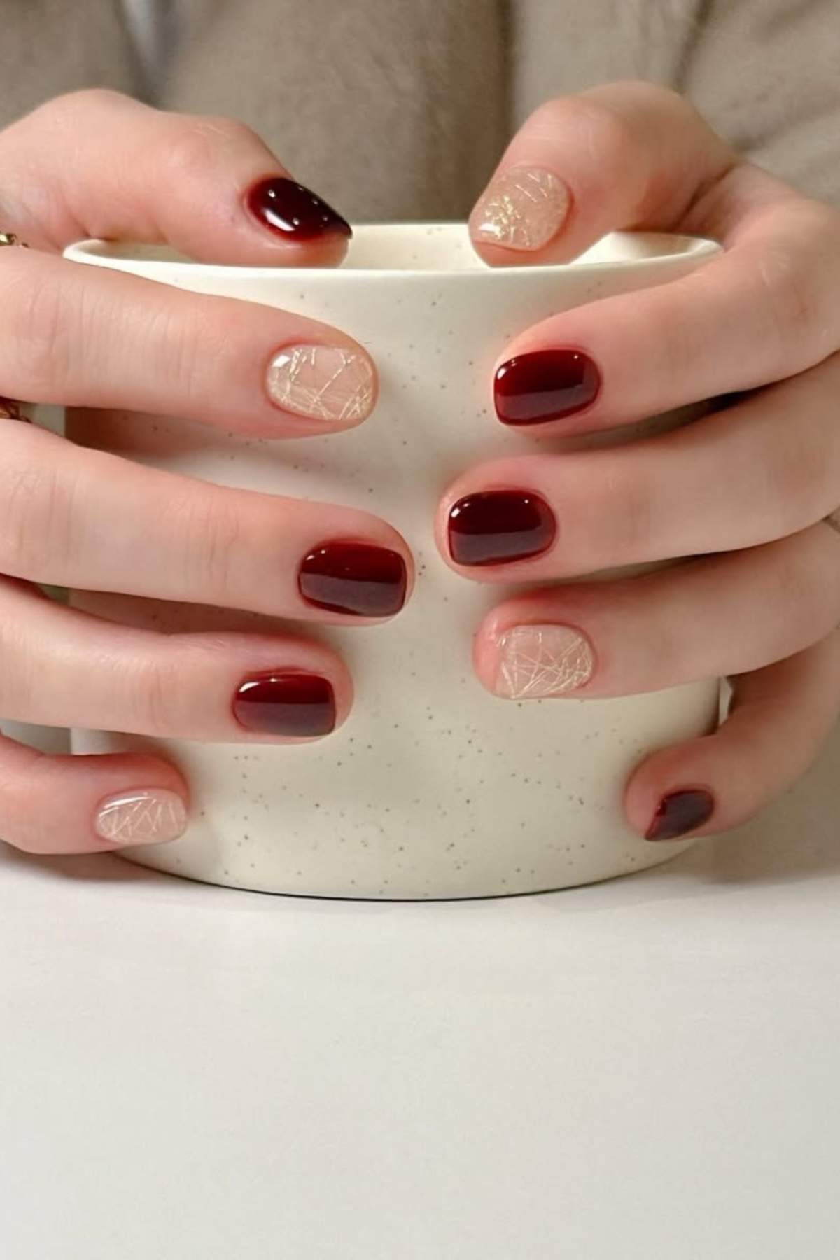 Hands holding a white speckled mug, nails painted burgundy with accent nails in nude and gold geometric designs.