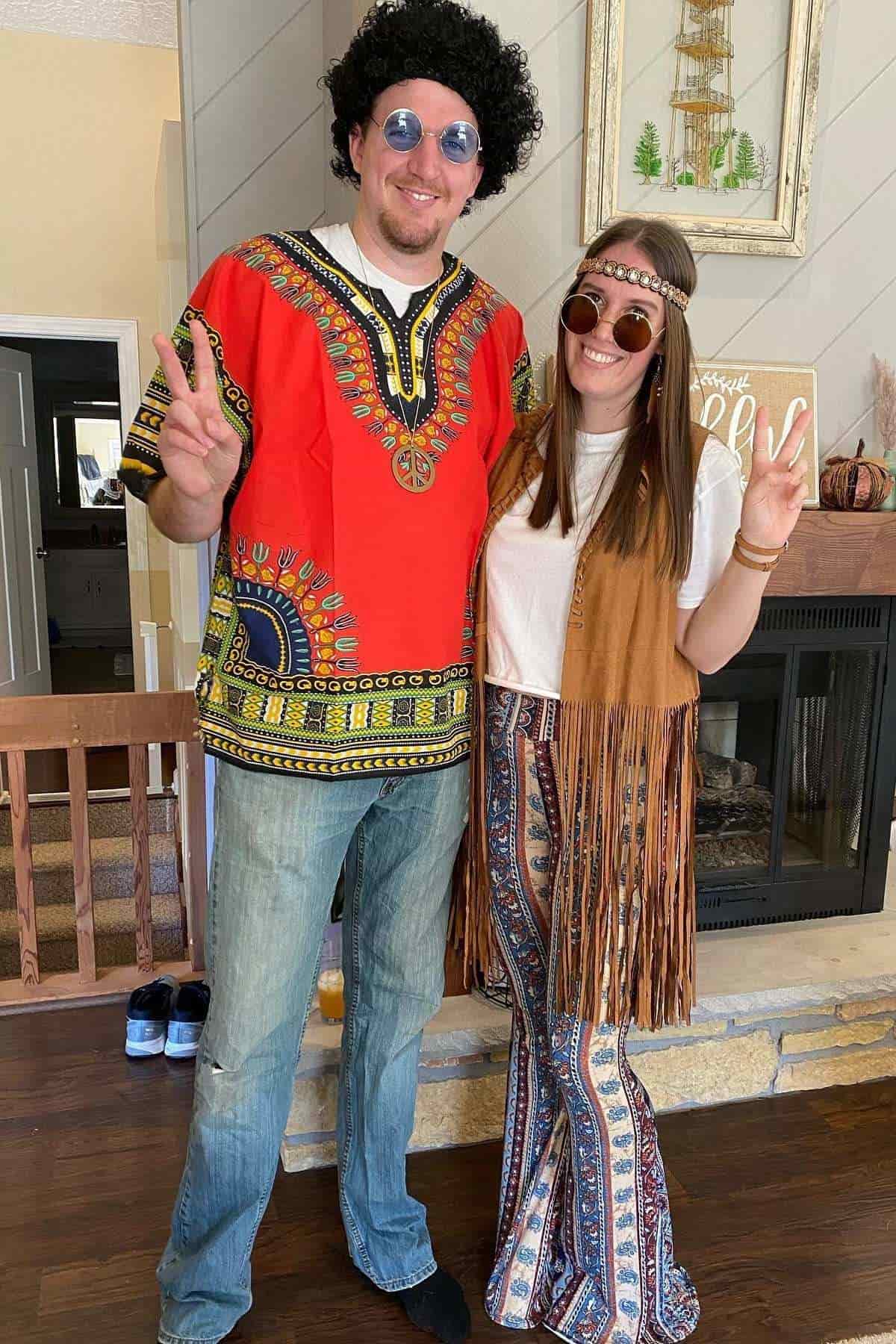 Two adults in retro 1970s costumes stand indoors, both making peace signs. The man wears a red dashiki and wig; the woman wears a fringe vest, patterned pants, and a headband.