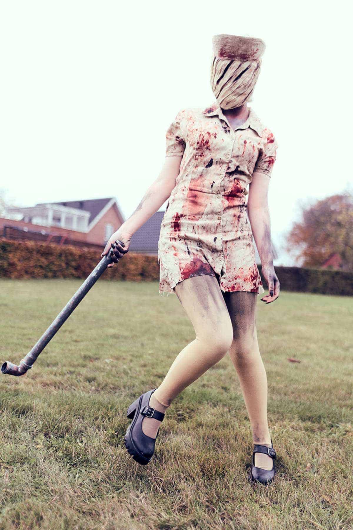 Person in a blood-stained nurse costume with a bandaged head, holding a metal pipe, stands on grass with houses and trees in the background.