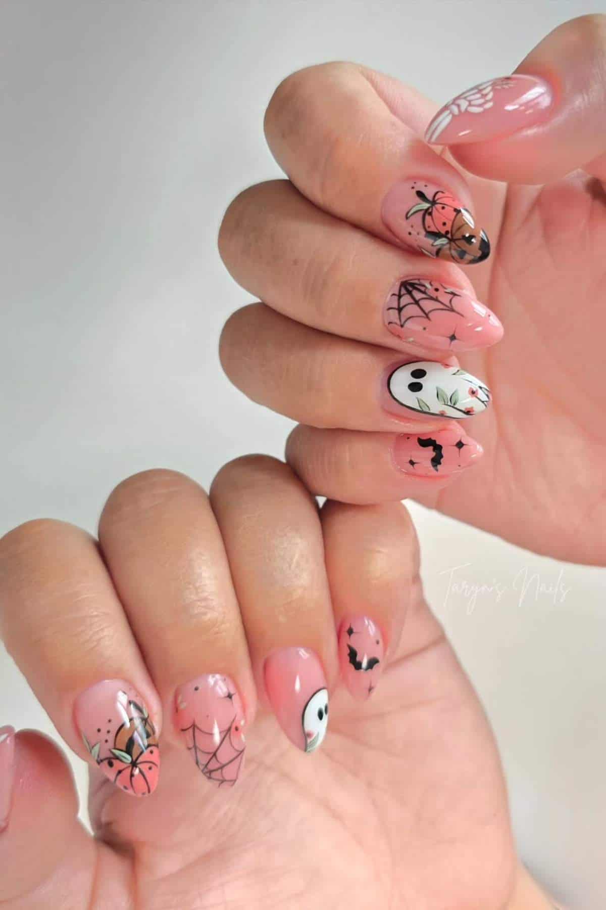 Hands with long, almond-shaped nails painted pink and decorated with Halloween designs, including spiders, spider webs, bats, and white ghosts.