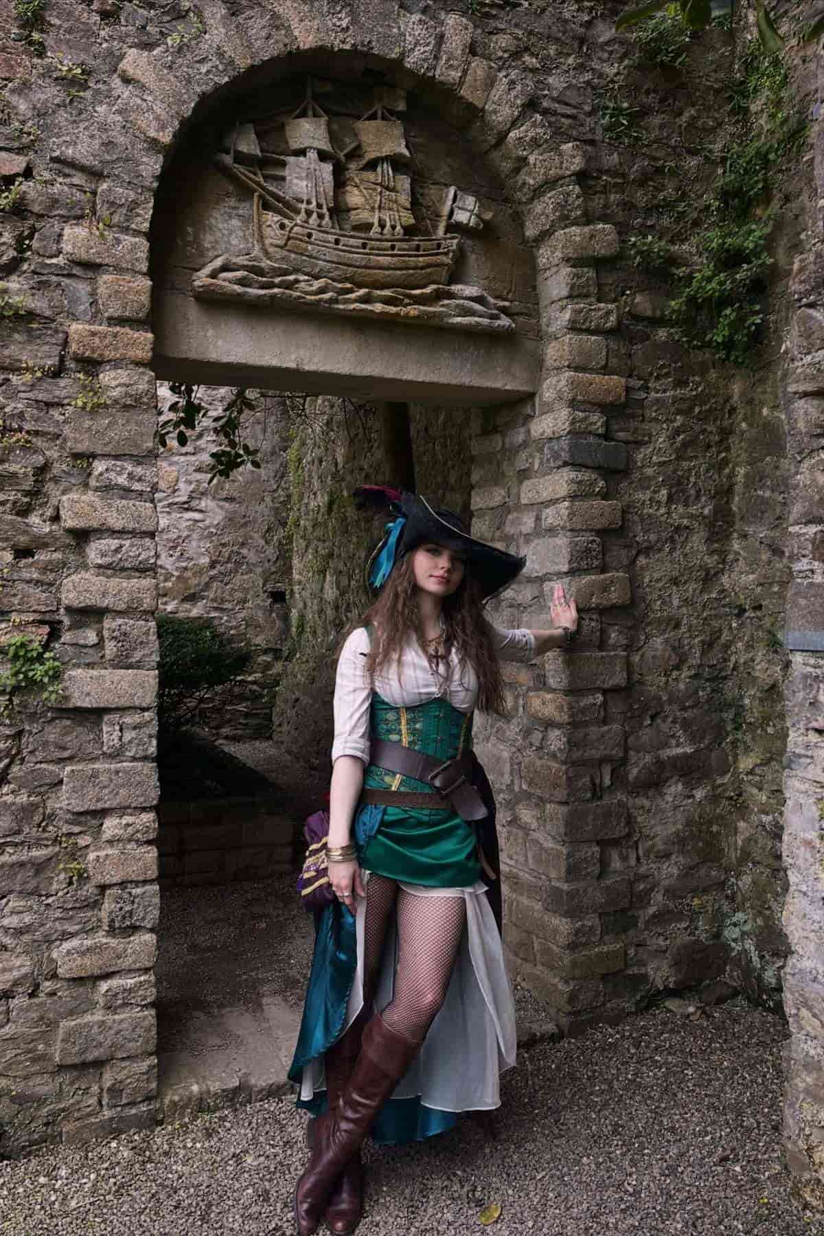 Person in a pirate costume stands by a stone archway with a ship carving above, surrounded by rough stone walls and greenery.