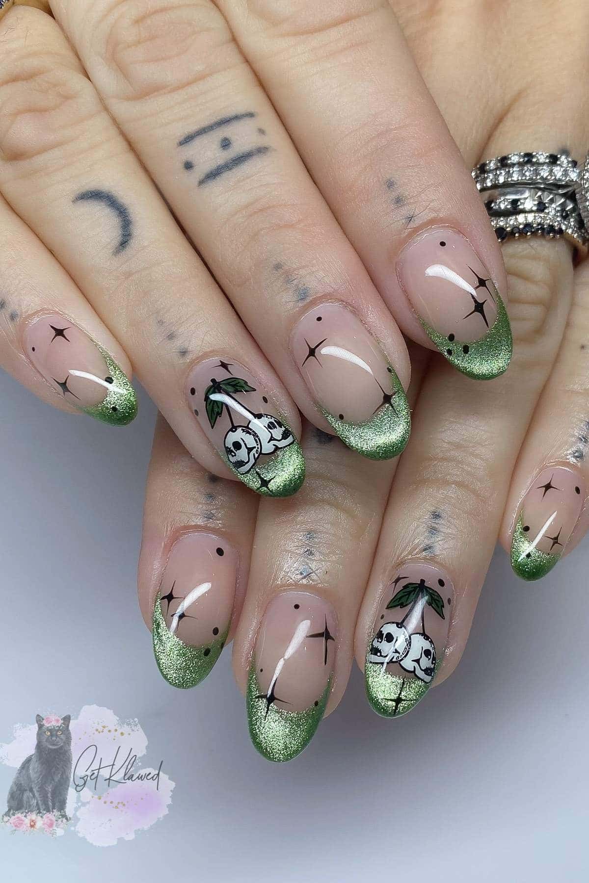 Hands with green glitter French tip nails, black star designs, and mushrooms with skulls on two nails; fingers have small tattoos.