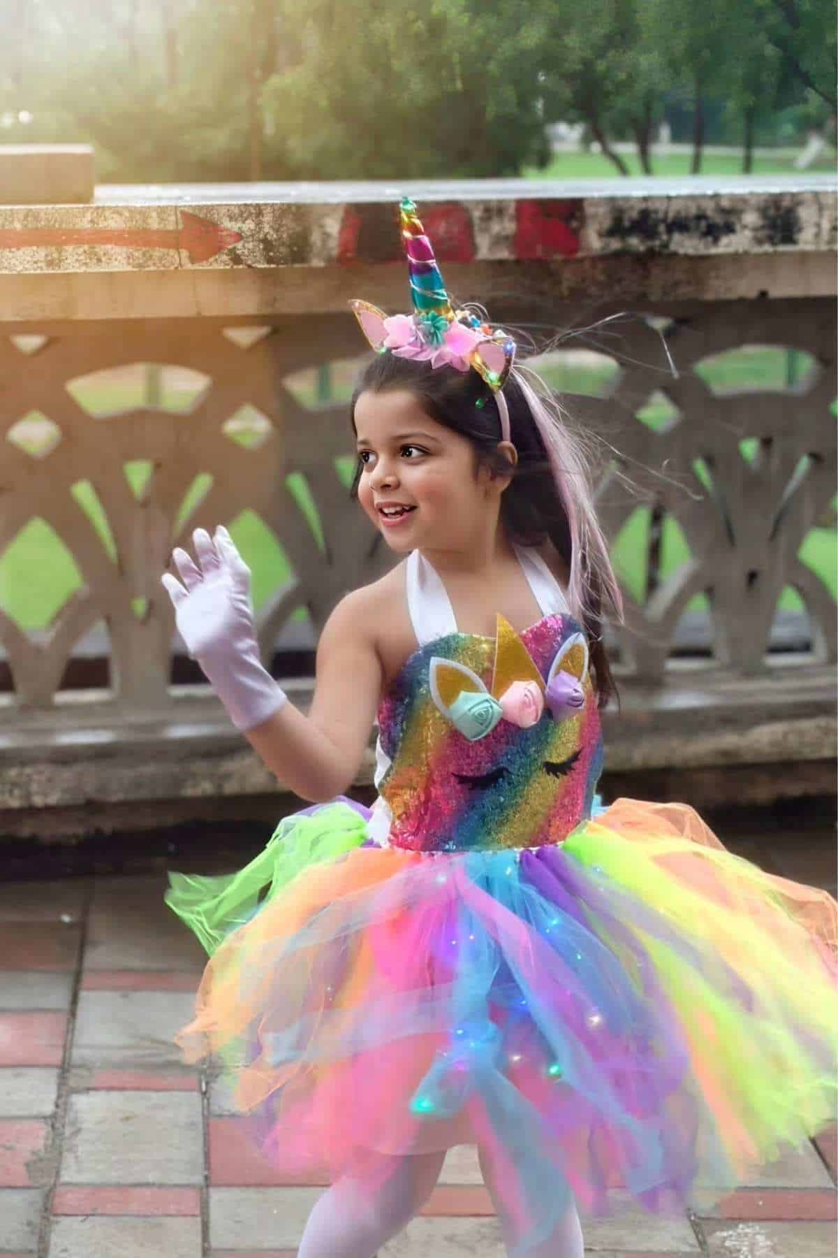 A young child wearing a colorful unicorn costume and gloves waves while standing outdoors on a tiled surface near a stone railing.