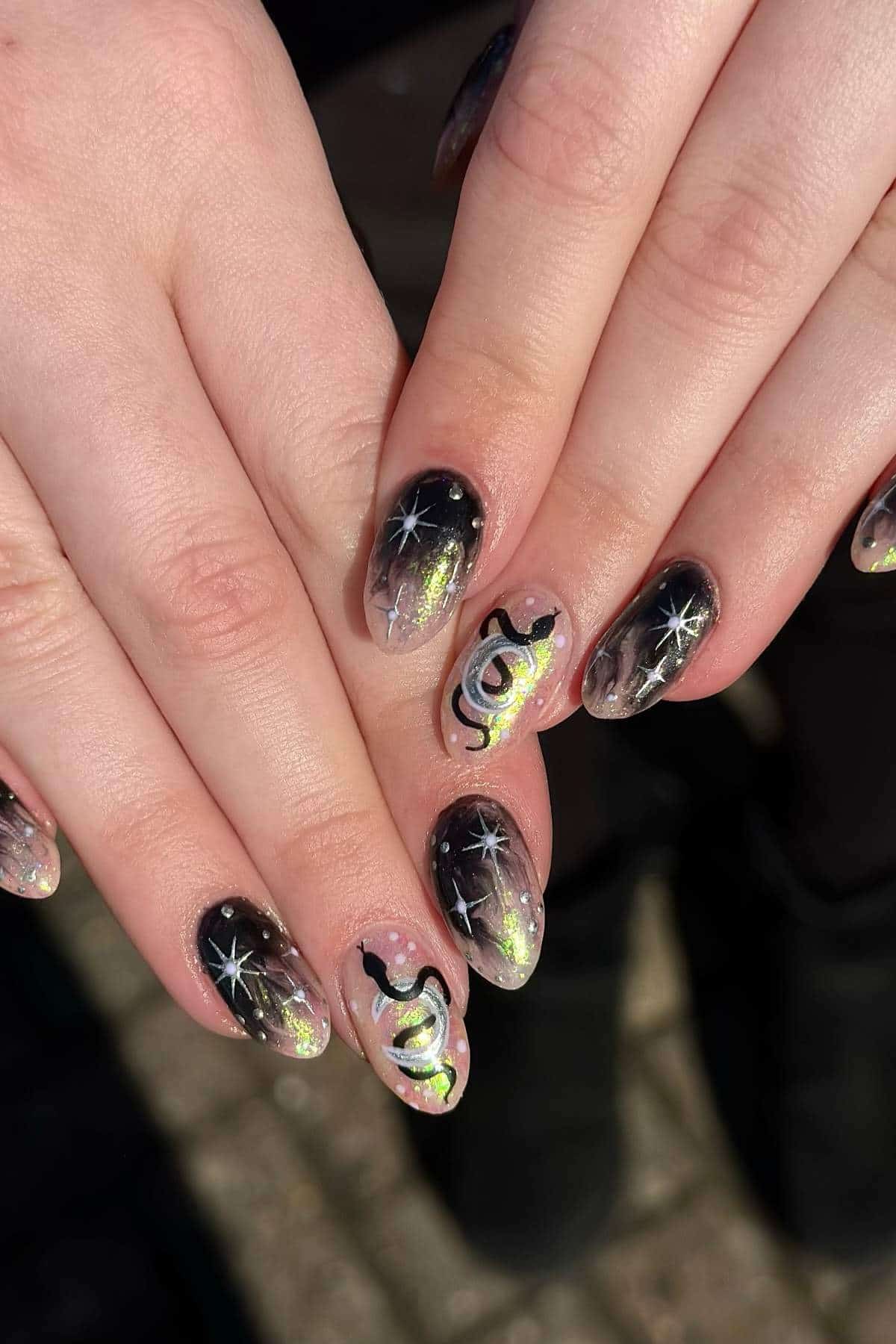 Hands with almond-shaped nails featuring black ombre, iridescent glitter, white star designs, and abstract black and neon yellow patterns on two accent nails.