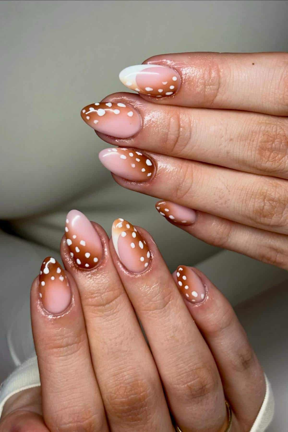 Hands with almond-shaped nails featuring a brown ombré design and white polka dots, with one accent nail having a white tip. Nails are manicured and neatly painted.