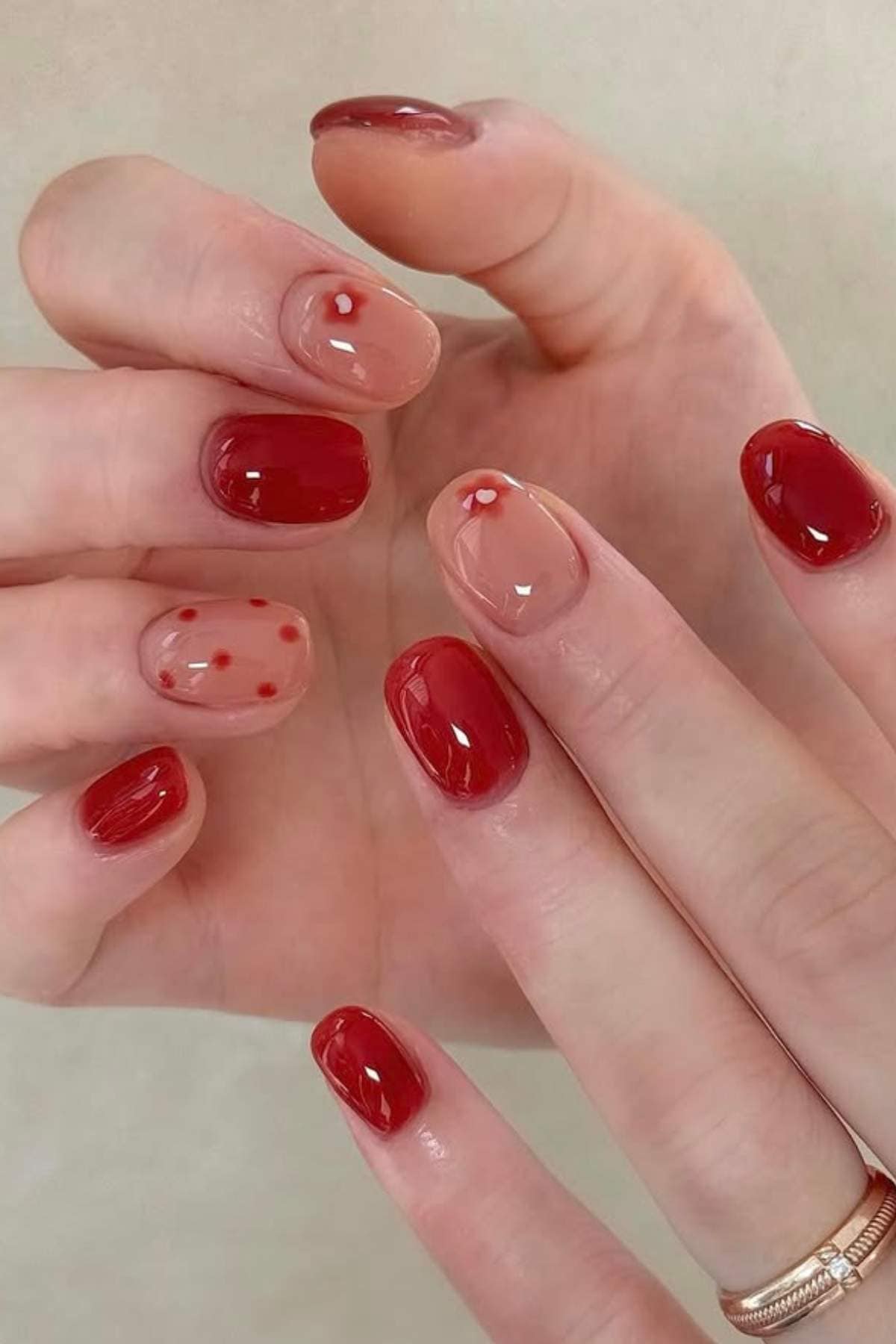 Close-up of hands with red and nude nails; some nails have small red dots or heart designs, while others are painted solid red.