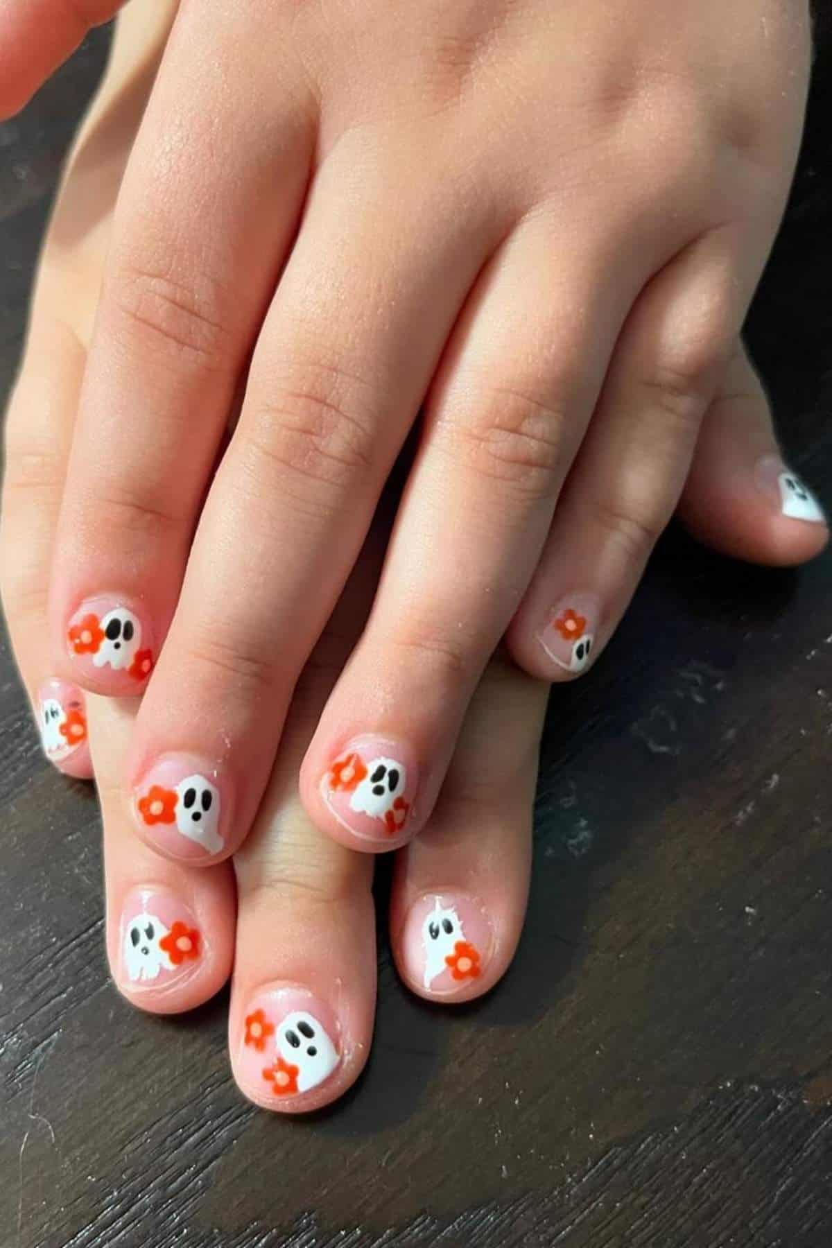 Fingernails with white ghost and orange flower designs are shown resting on a dark surface.