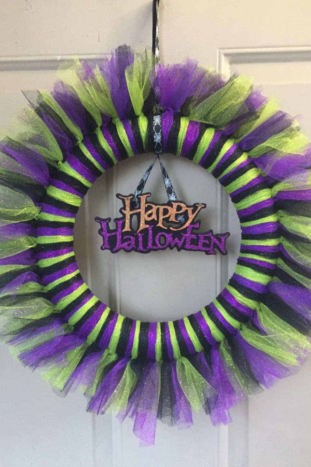 A wreath with purple, green, and black tulle hangs on a door, featuring a sign in the center that reads “Happy Halloween” in glittery letters.