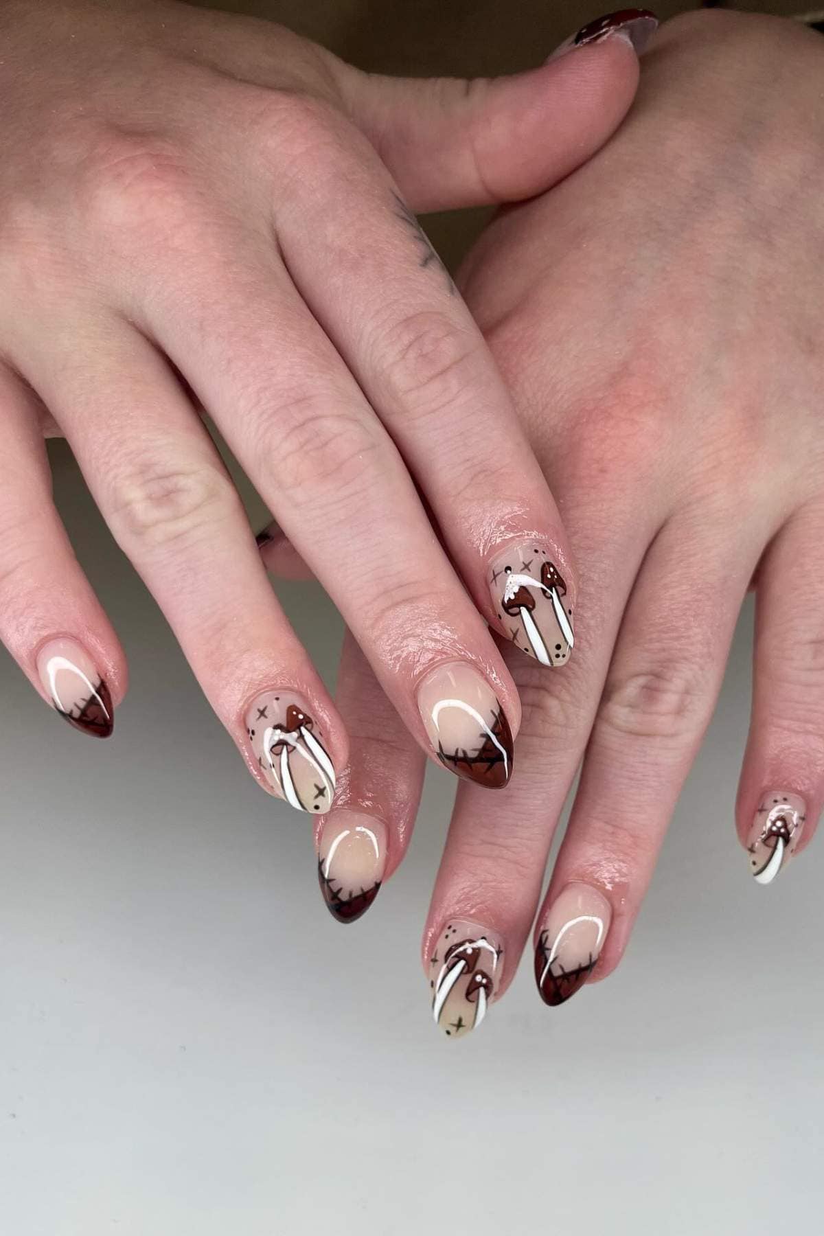 Two hands with almond-shaped nails featuring a brown and beige abstract design with curved lines and dots on a white surface.