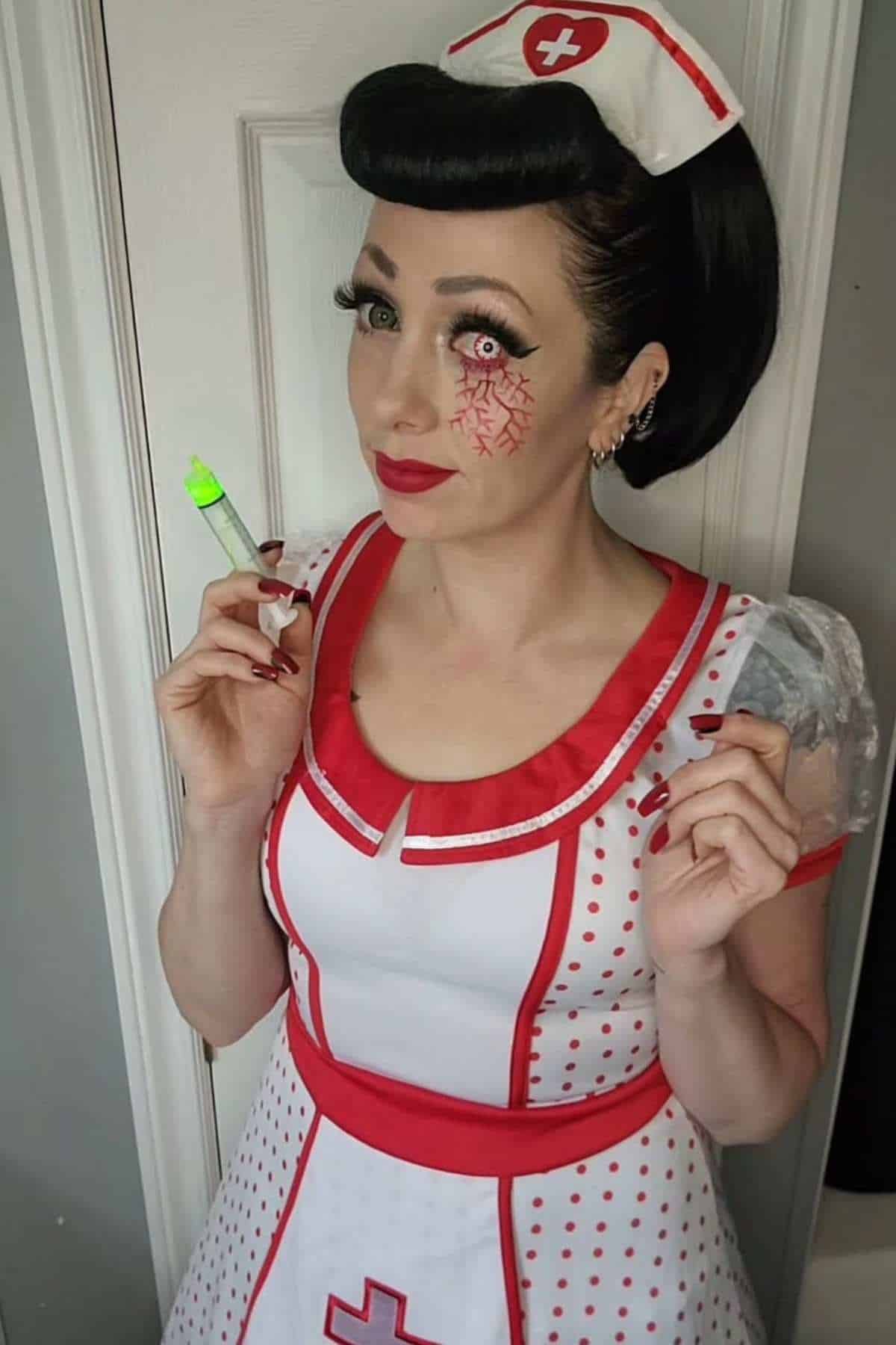 Person in a retro nurse costume with red accents holds a green-capped syringe and has red vein makeup extending from one eye, standing indoors near a white door.