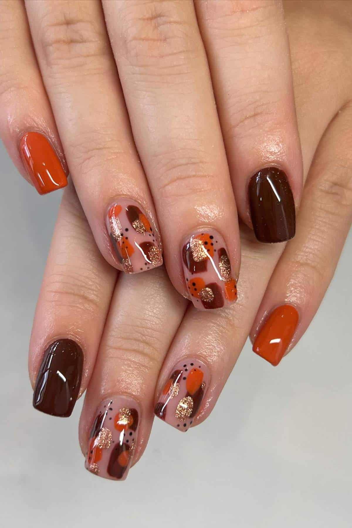 Hands with short nails featuring a fall-inspired manicure in shades of brown, orange, and glitter, with abstract patterns and solid accent nails.