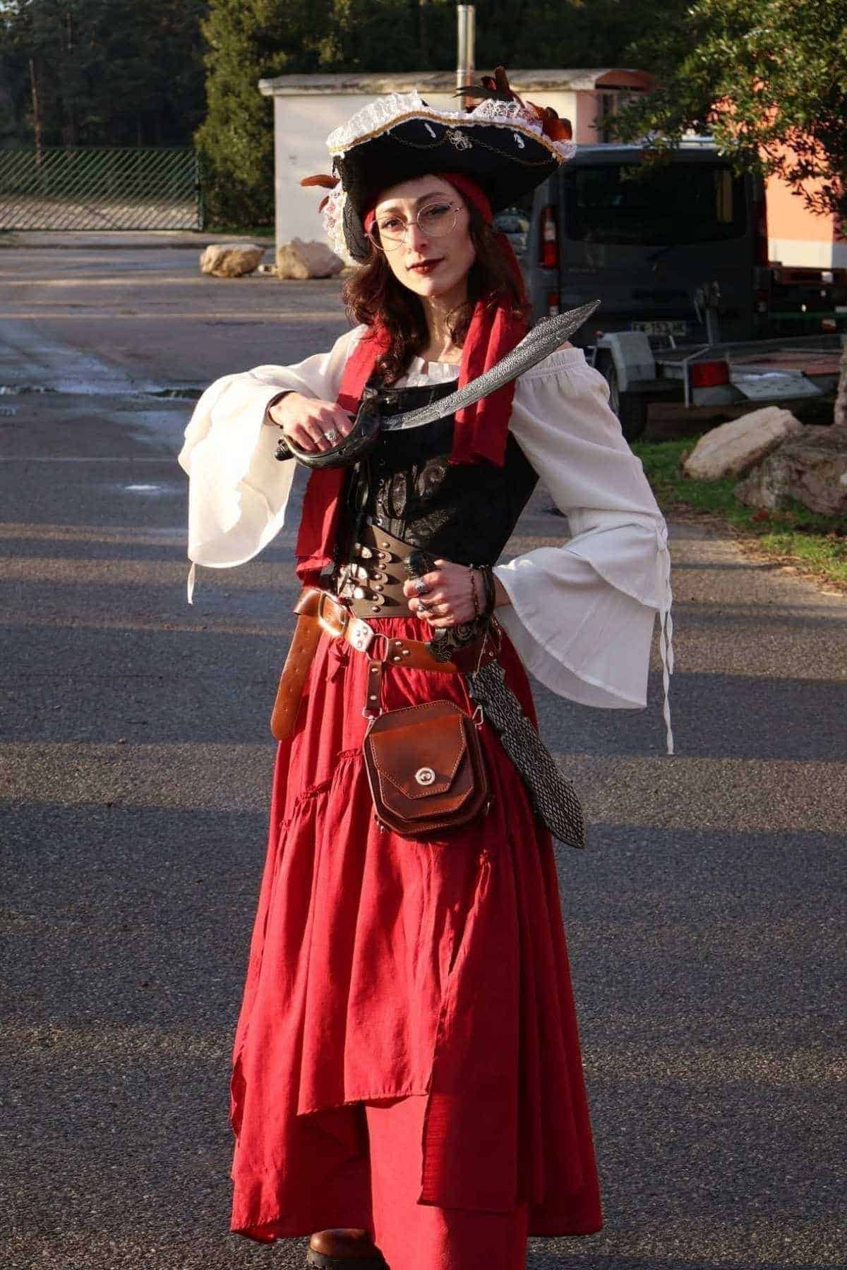 Person dressed in a pirate costume, wearing a hat and red skirt, holding a sword, standing outdoors on a paved surface.