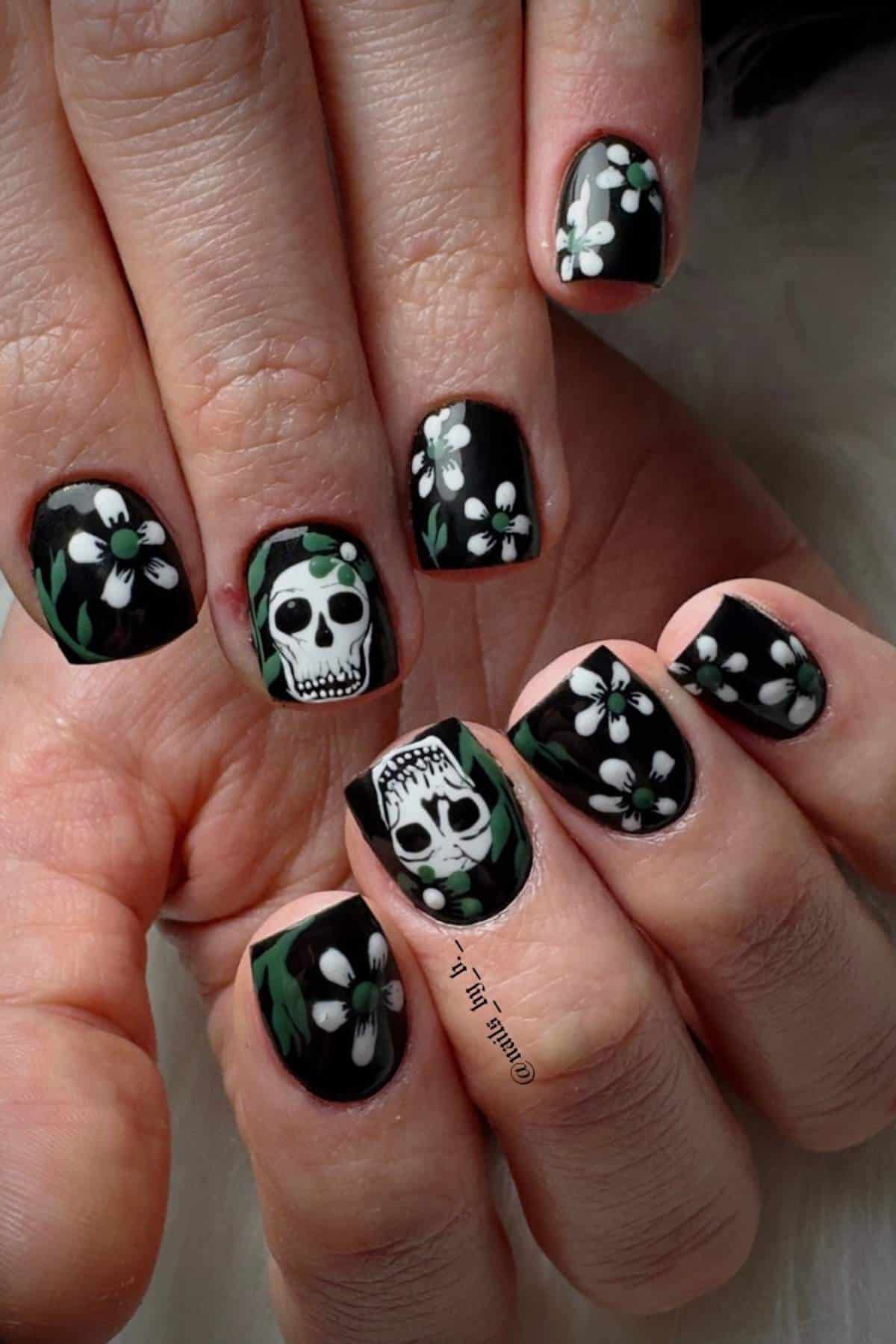 Hands with black nail polish featuring white flowers and two skull designs on the middle and ring fingernails.